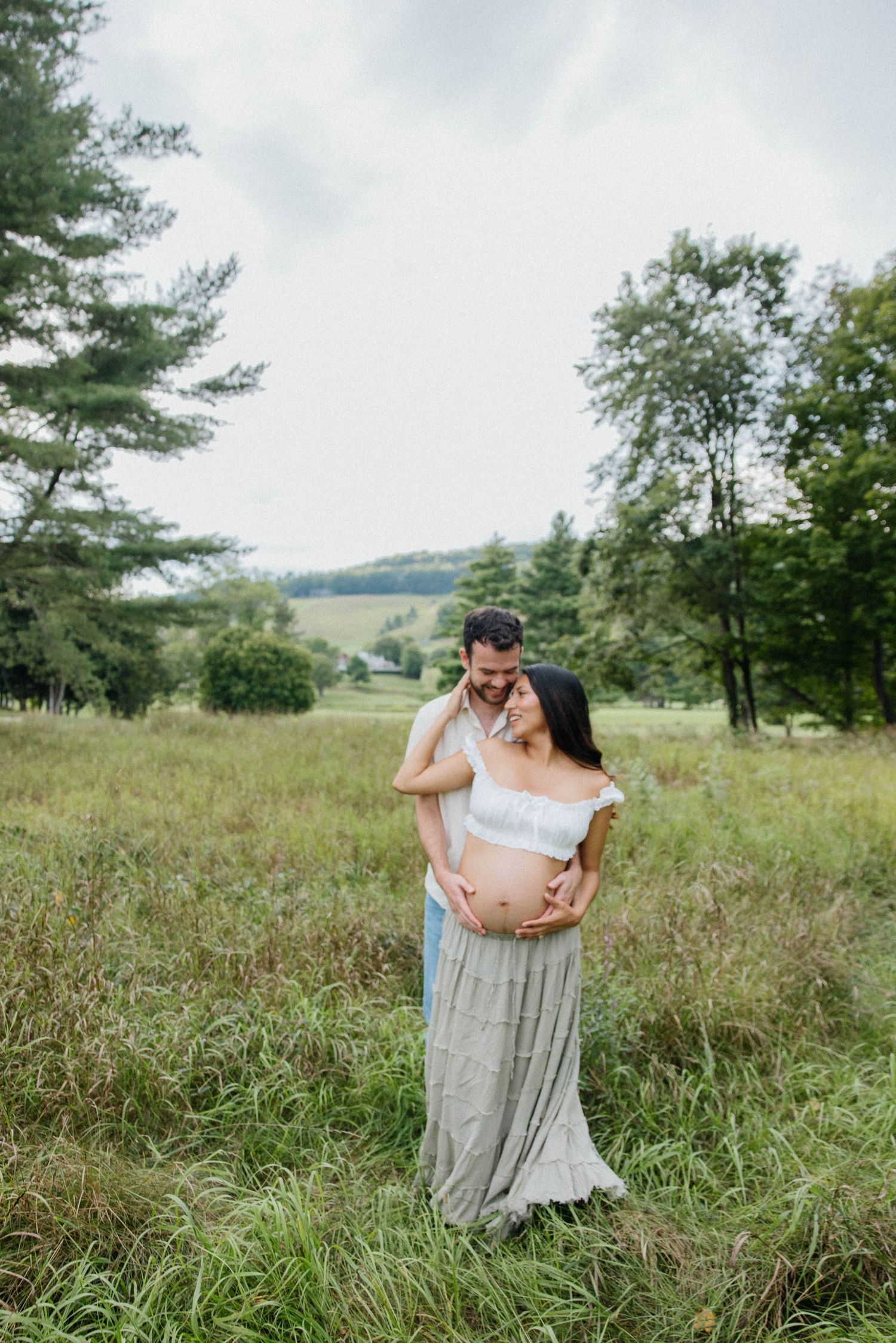 A couple shares a tender moment in a lush green field, cradling a baby bump during an outdoor maternity photo session.
