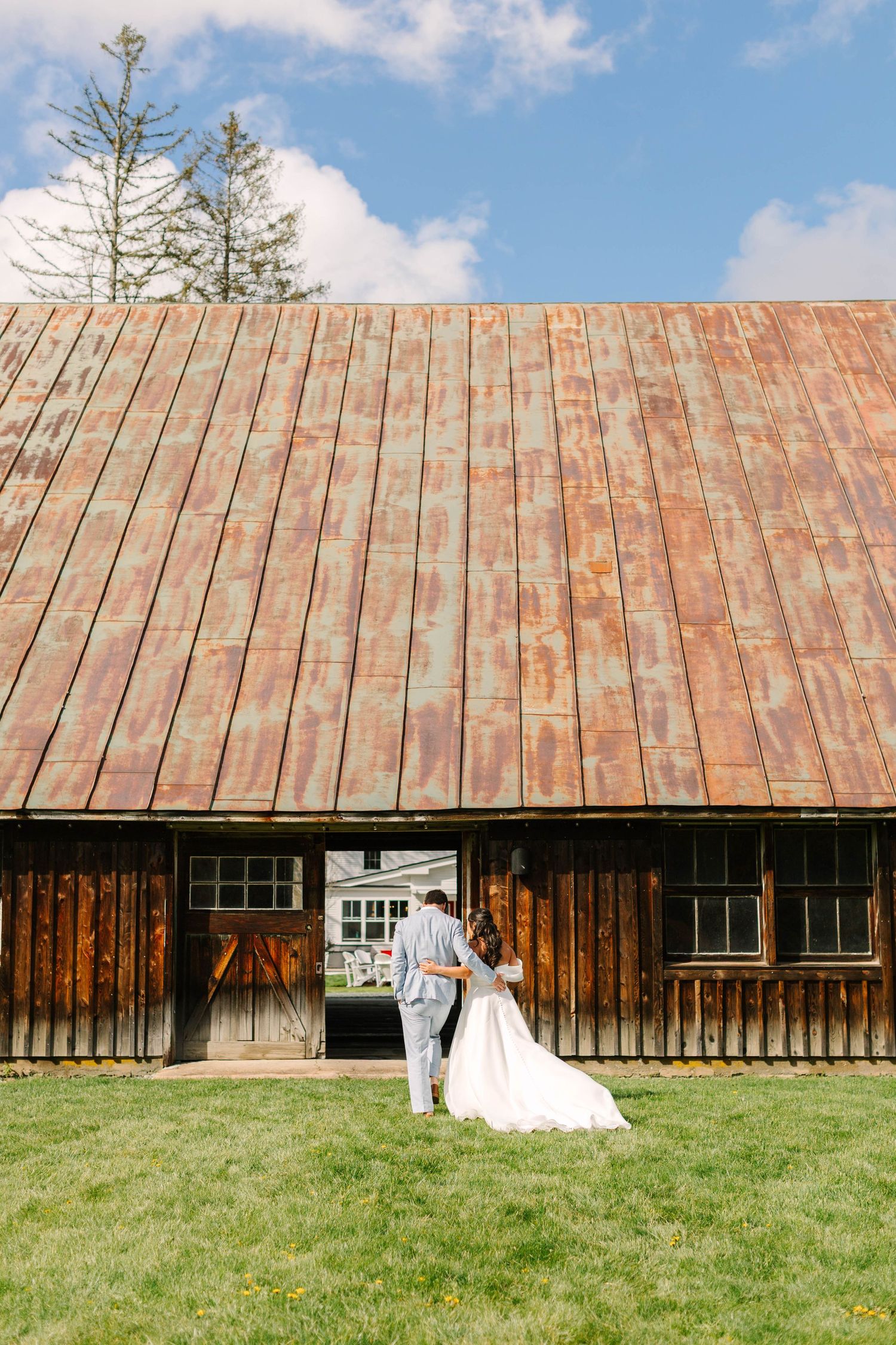 Bride and groom walk hand-in-hand toward rustic wooden barn with copper roof on wedding day.