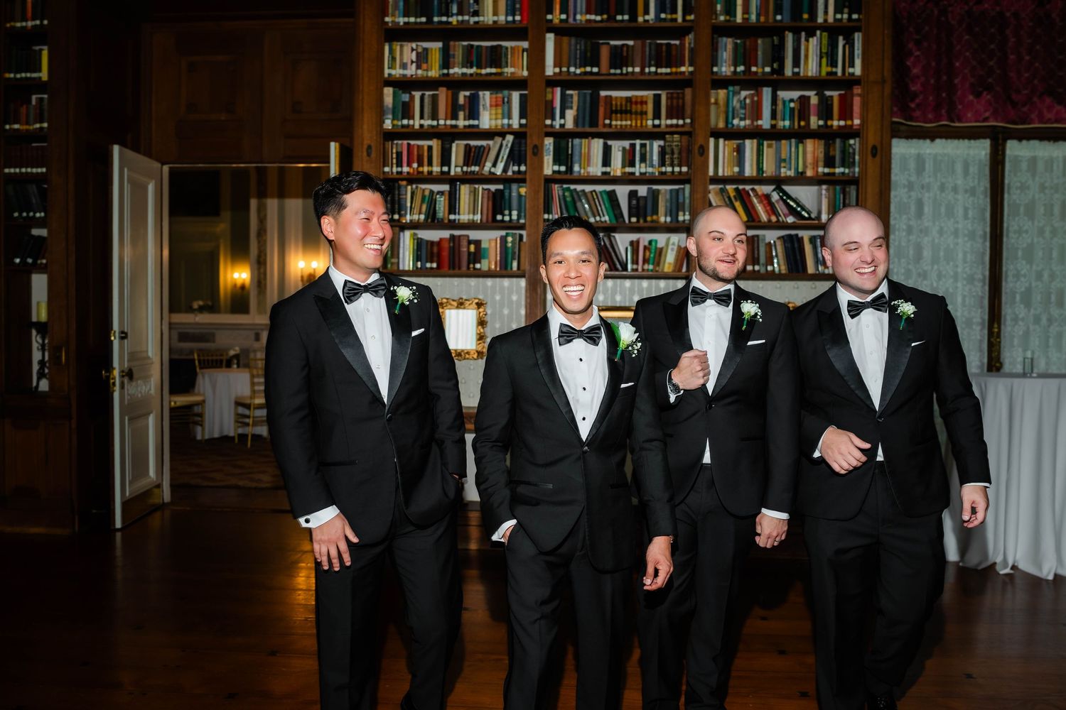 Four groomsmen in black tuxedos smiling together in an elegant library setting.