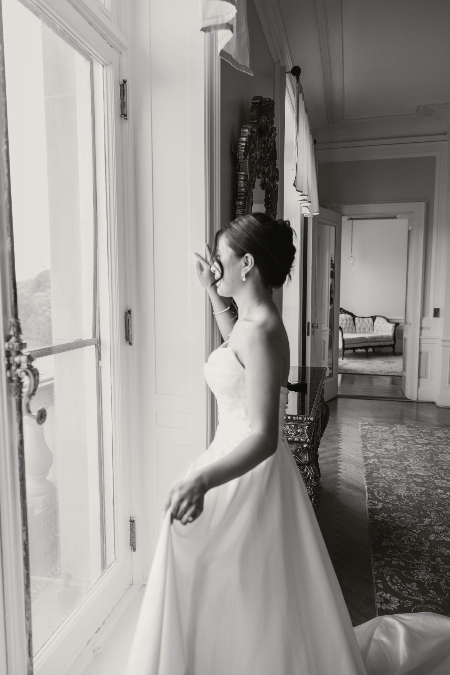 Bride in strapless white gown gazes out window in elegant black and white wedding portrait.
