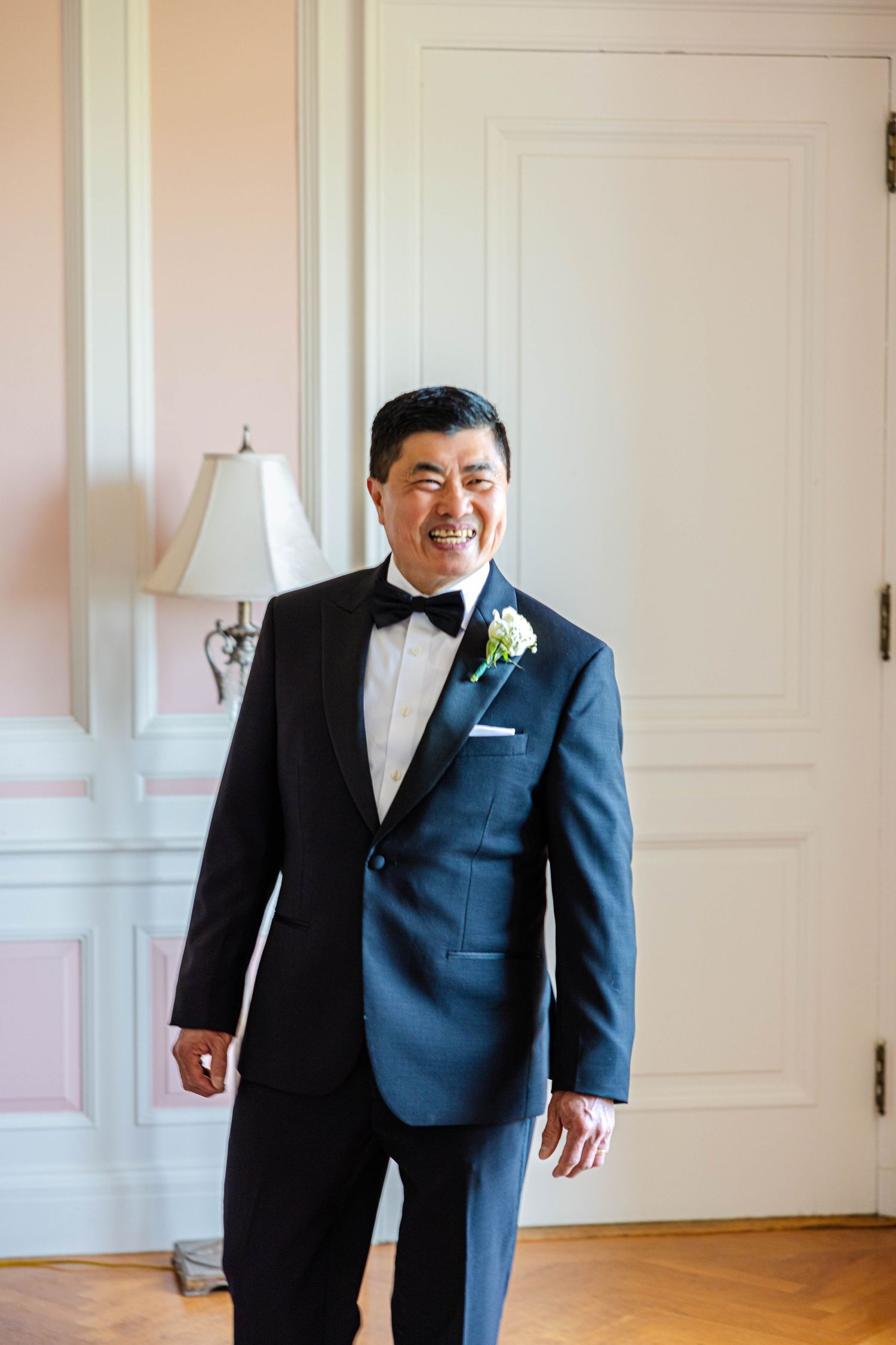 Smiling groom in navy tuxedo with bow tie and white boutonniere standing in elegant room.