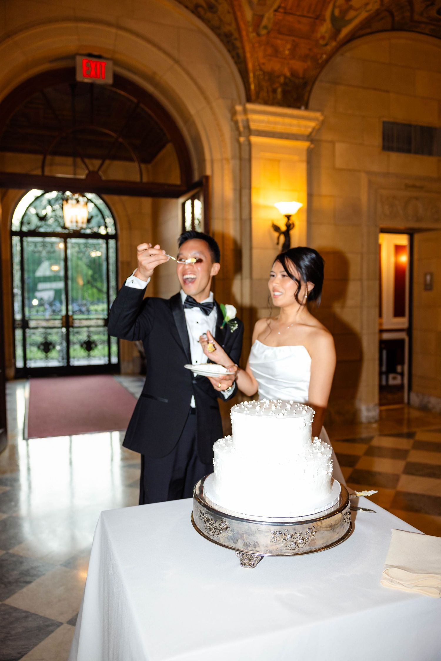 Bride and groom laugh while cutting elegant white tiered wedding cake in ornate ballroom venue.