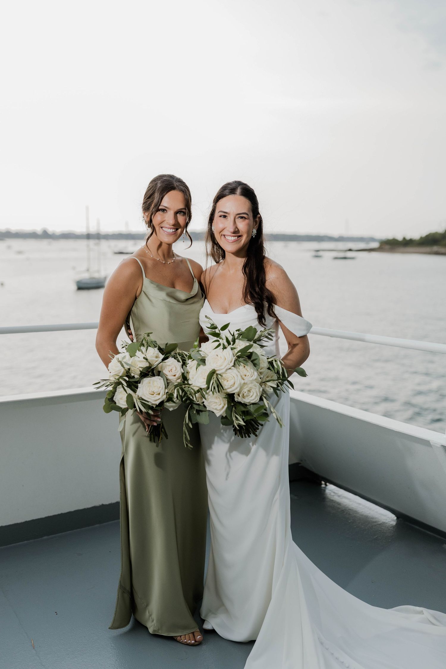 Bride and bridesmaid in sage green gown pose with white rose bouquets on waterfront boat venue.