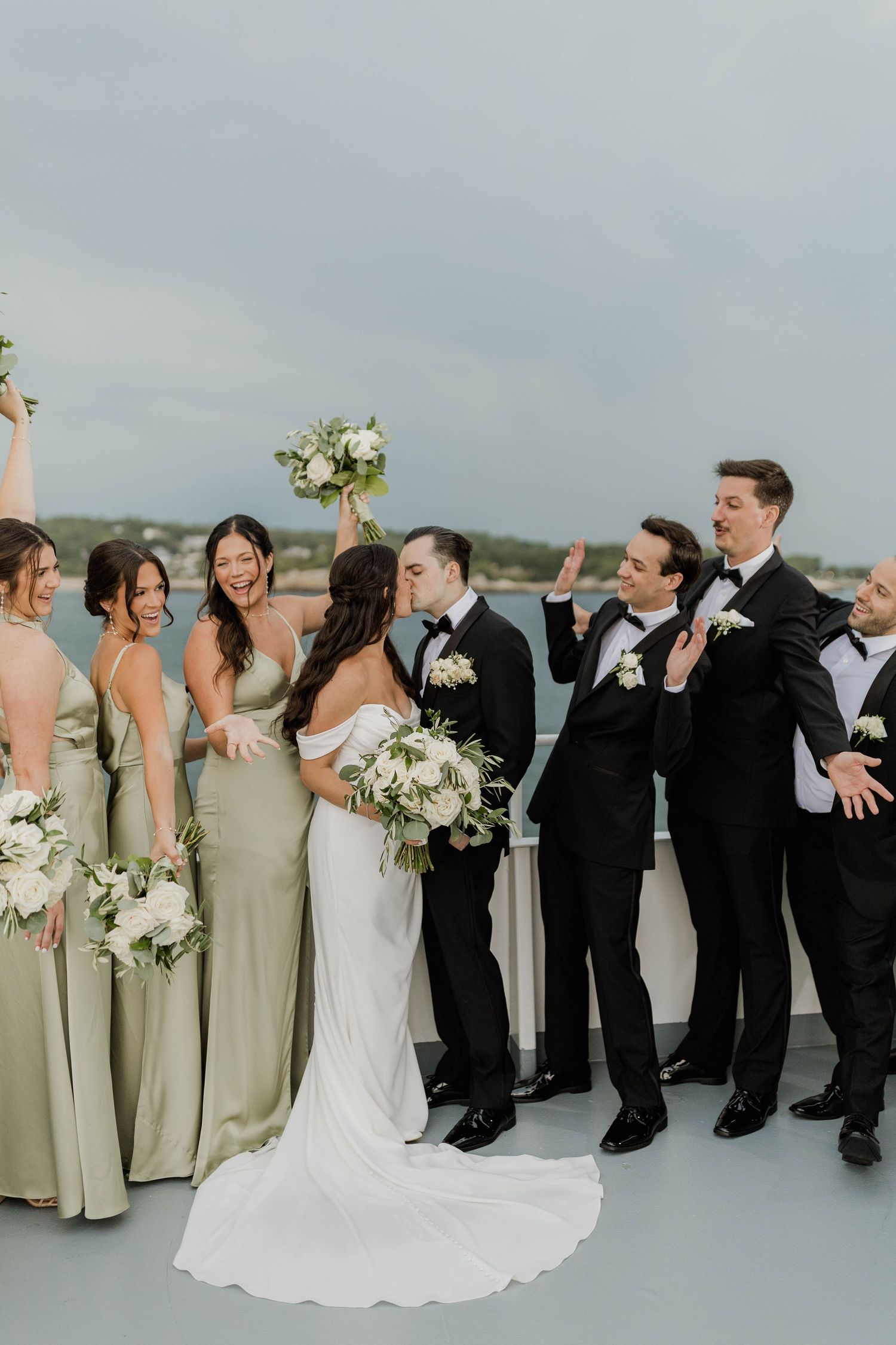 Wedding party on a boat deck, bride and groom kissing, bridesmaids in sage green gowns, groomsmen in black tuxedos.