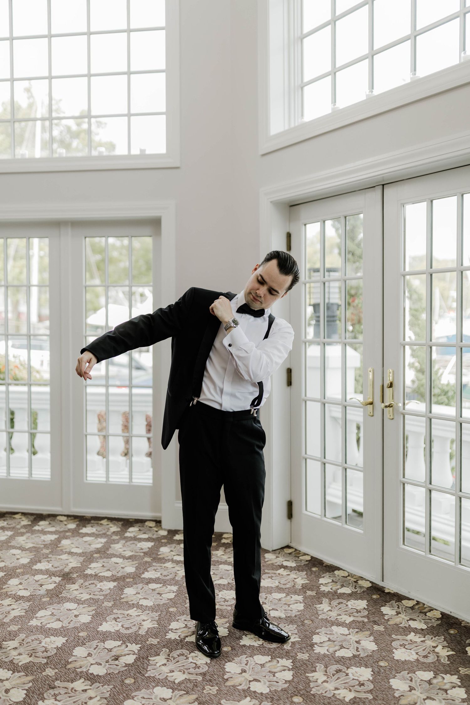 Dapper groom in a black tuxedo and bow tie adjusting his jacket in a bright windowed room.
