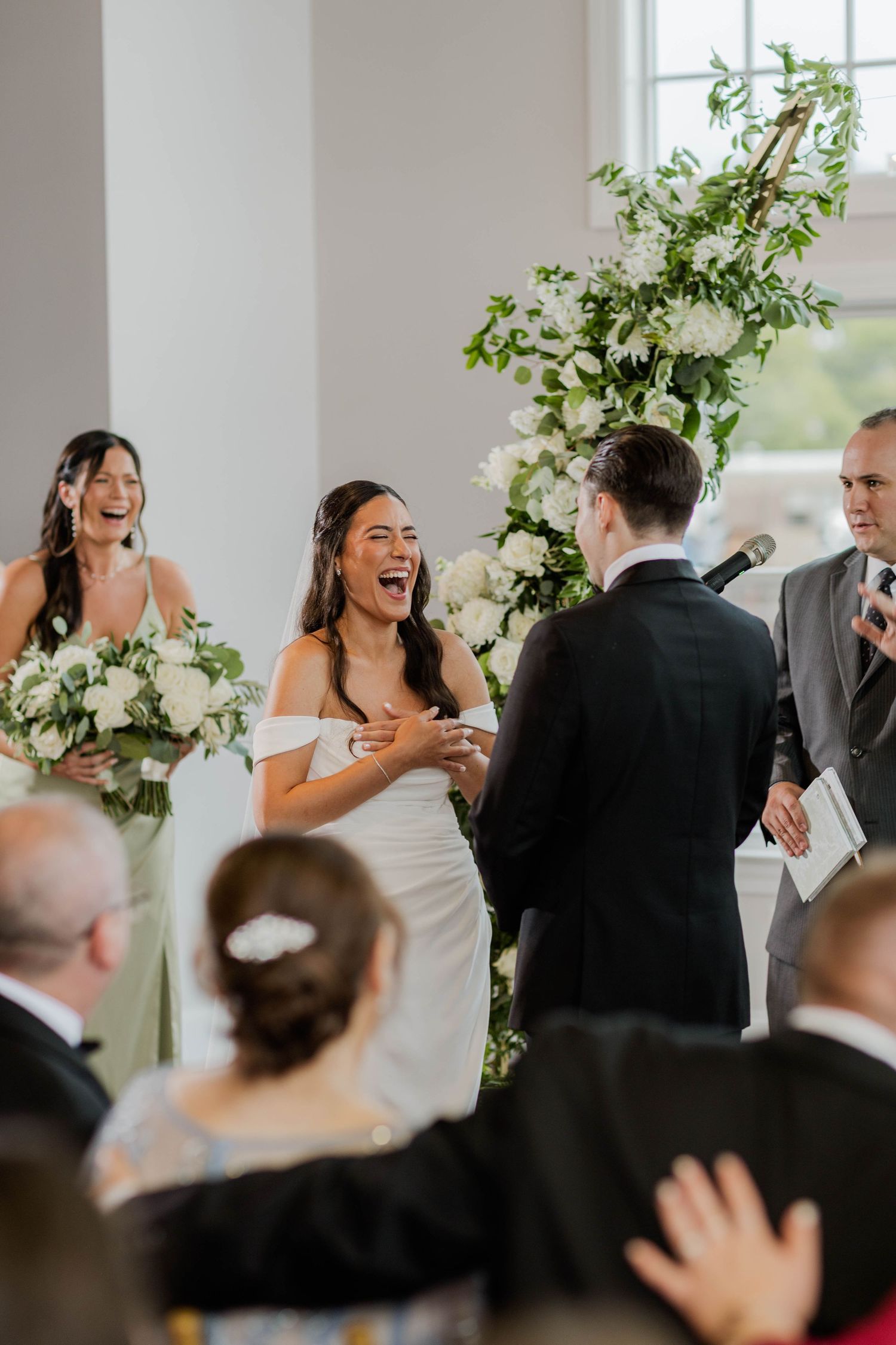 Joyful bride laughing at the altar with bridesmaids and floral arch during indoor wedding ceremony.