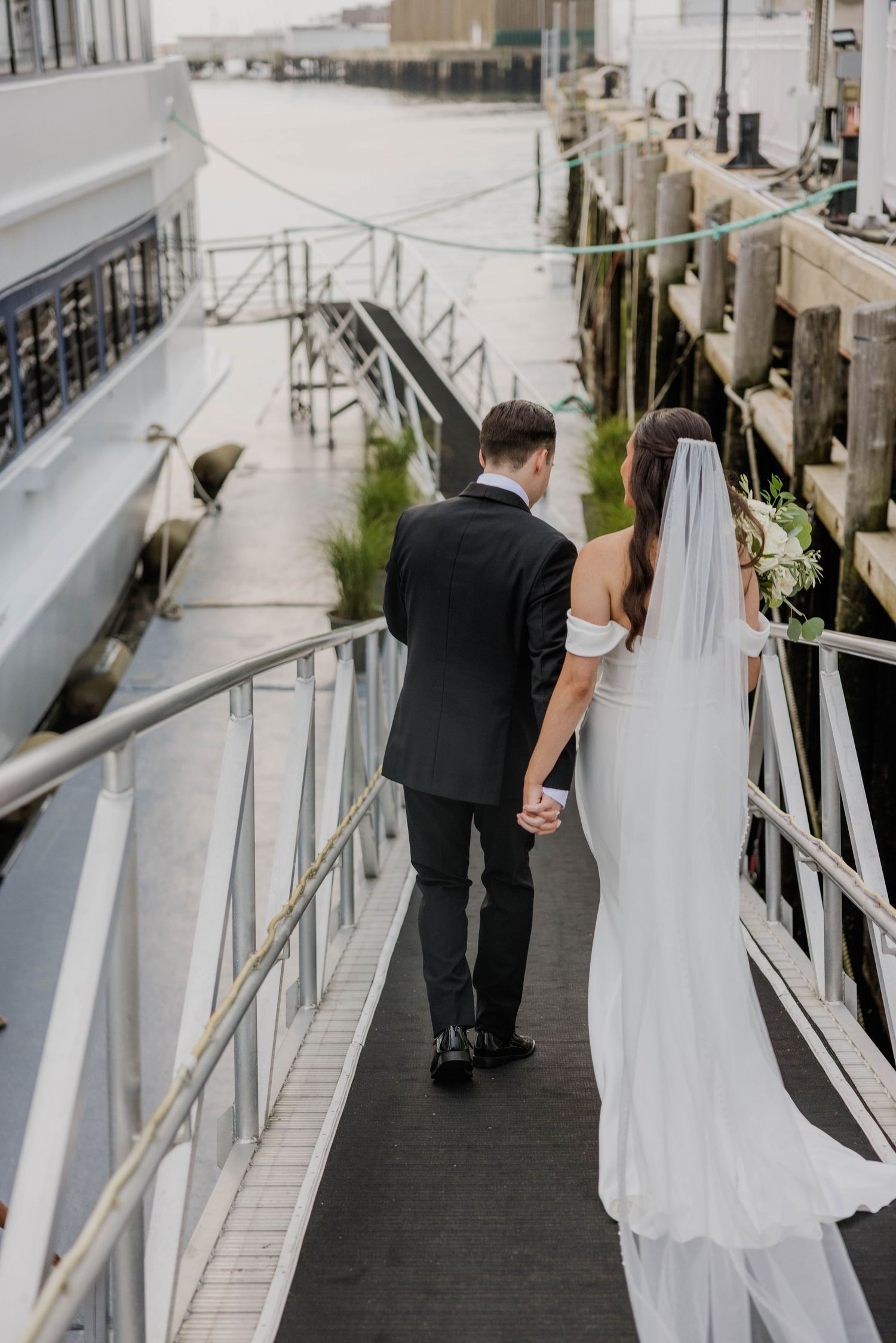 Bride and groom walking hand in hand along a waterfront pier, bride wearing a flowing white gown with veil.