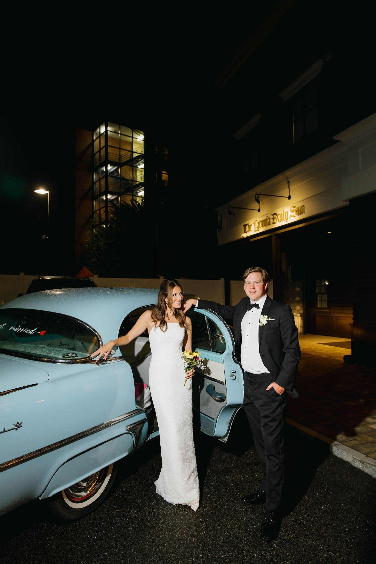 Bride and groom pose beside a vintage light blue car at night outside an elegant venue.