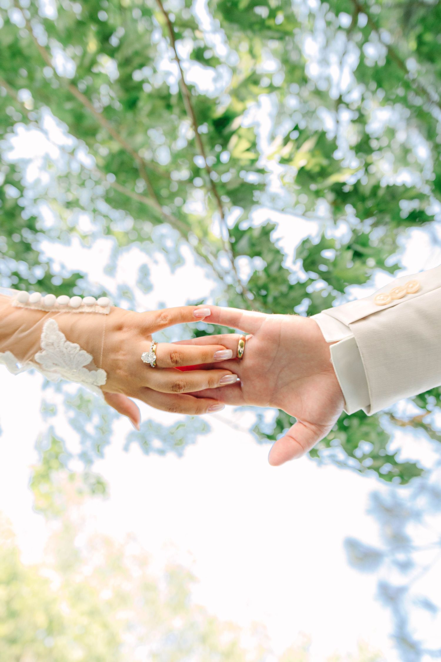 Close-up of bride and groom holding hands showing diamond engagement ring under green trees.