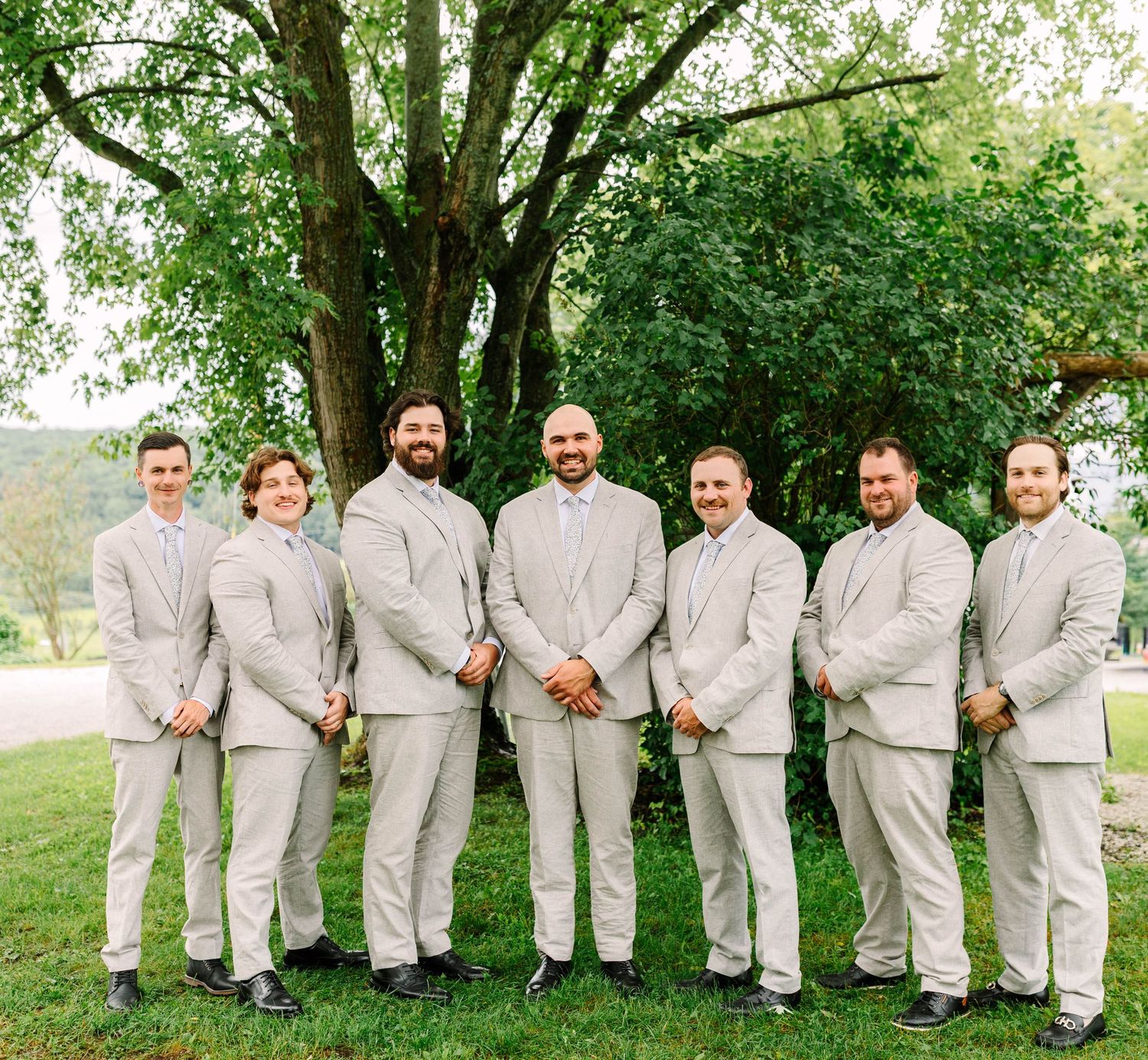 Groom and groomsmen pose in matching light gray suits under a large tree at an outdoor wedding.