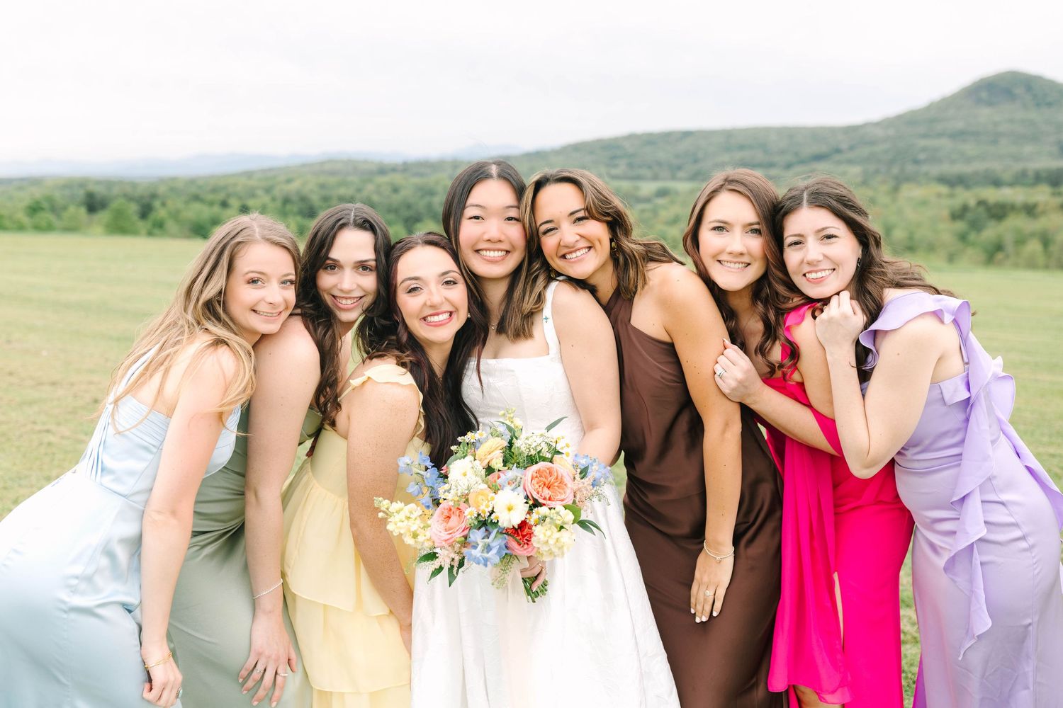 Smiling bride holding colorful bouquet poses with six bridesmaids in mismatched colorful dresses on a green field.