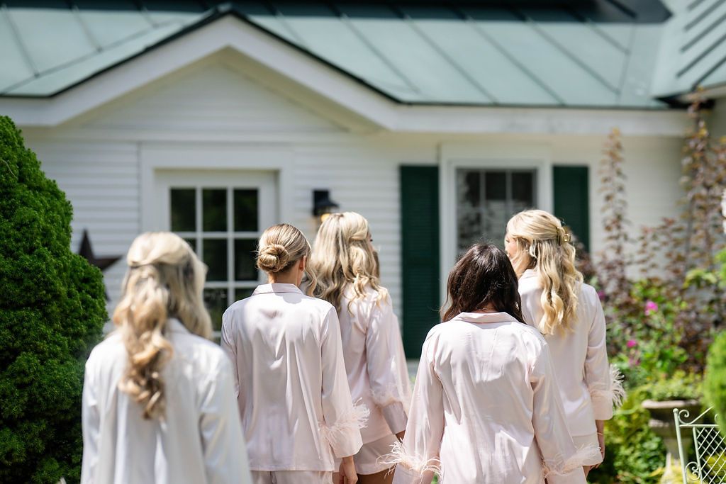 Bridesmaids in matching white robes walk toward a charming white house with green trim surrounded by lush gardens.