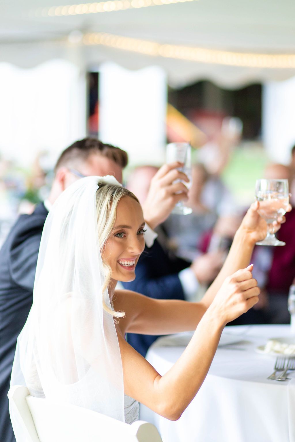 Smiling bride in white veil raises champagne glass during wedding reception toast with guests in background.