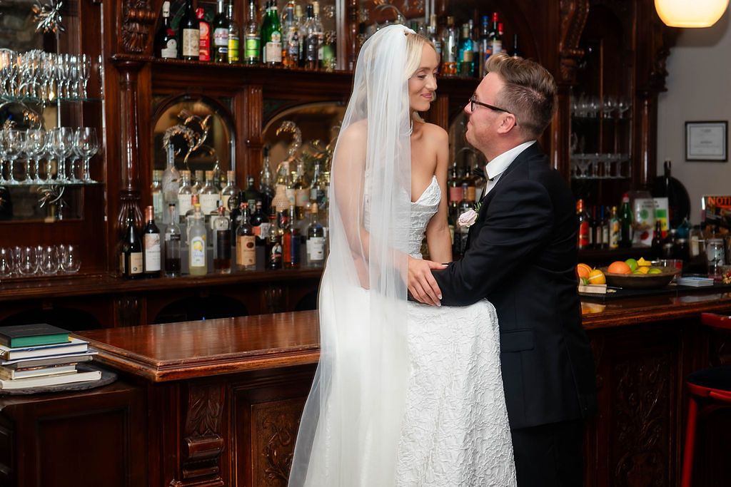 Bride and groom sharing romantic moment at vintage wooden bar, bride wearing lace wedding gown and veil.