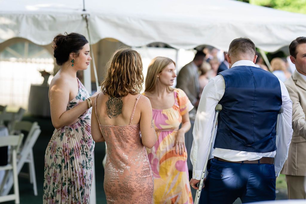 Elegantly dressed wedding guests socialize under a white tent, including a man in a navy suit at an outdoor reception.
