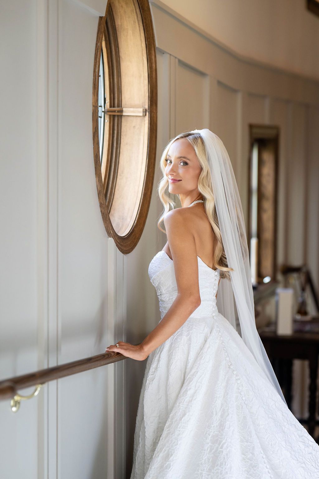 Smiling bride in strapless lace wedding dress and veil stands by railing near decorative round mirror.