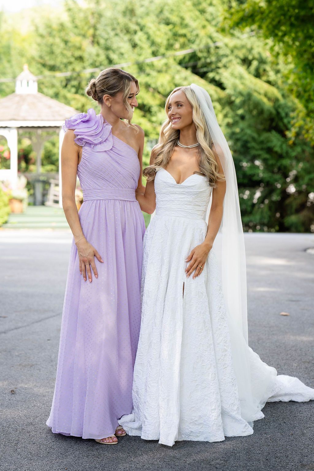 Bride in white lace gown with veil stands beside bridesmaid in lavender one-shoulder dress at outdoor wedding venue.
