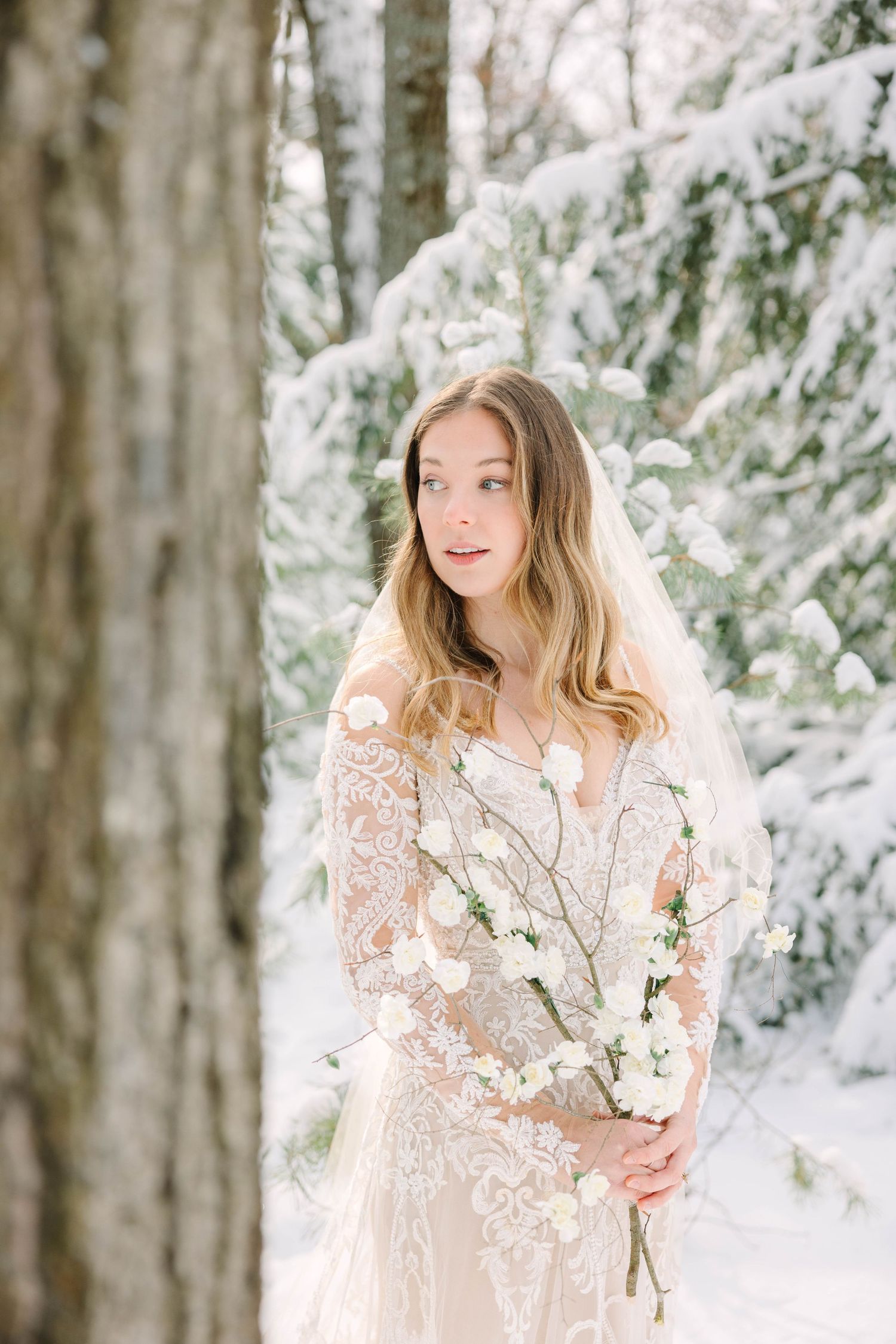 Elegant bride in lace wedding dress with veil posing in snow-covered winter forest.