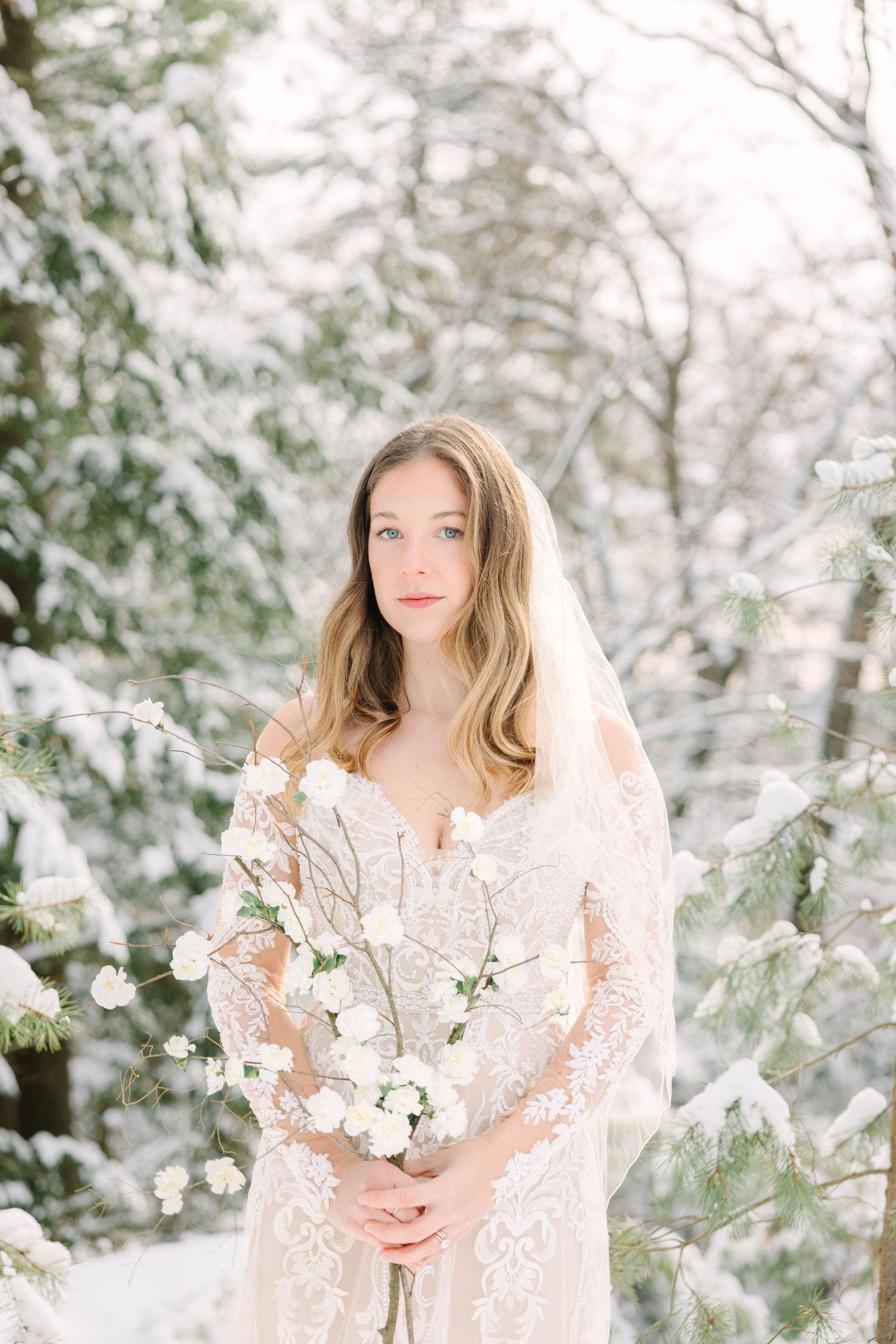 Bride in lace gown and veil holds white floral bouquet among snow-covered pine trees in a winter wonderland setting.