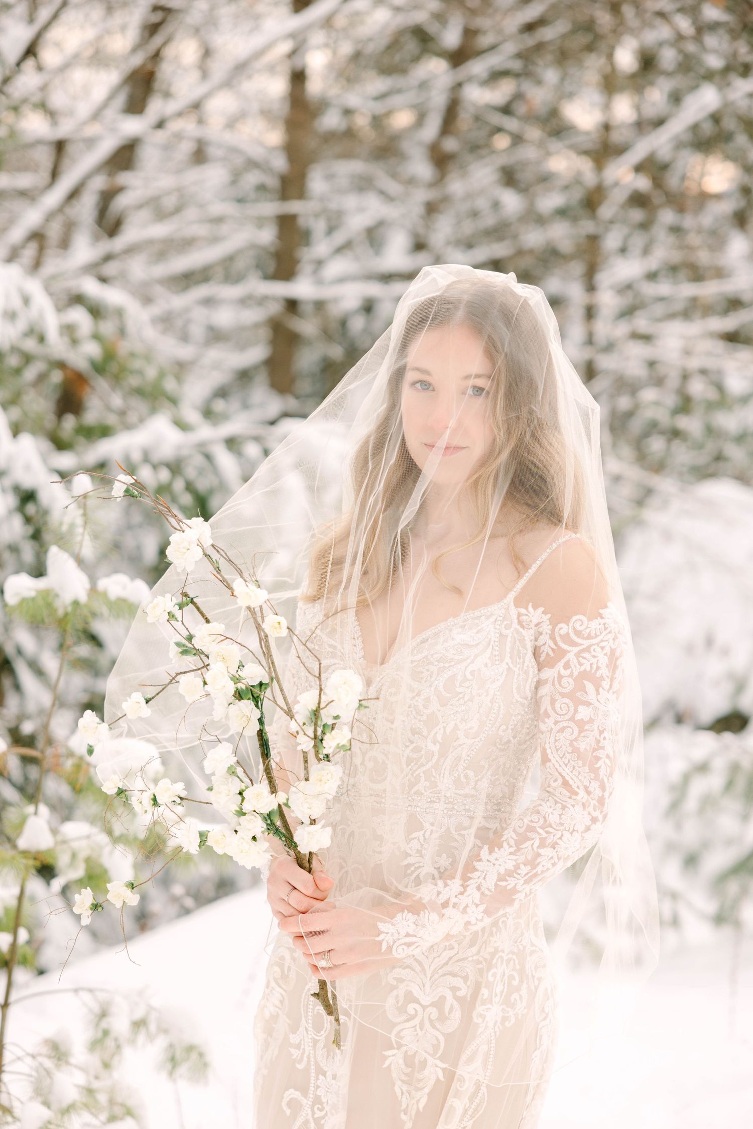 Bride in lace off-shoulder gown covered by veil holds white blossoms amid a glowing snowy winter landscape.