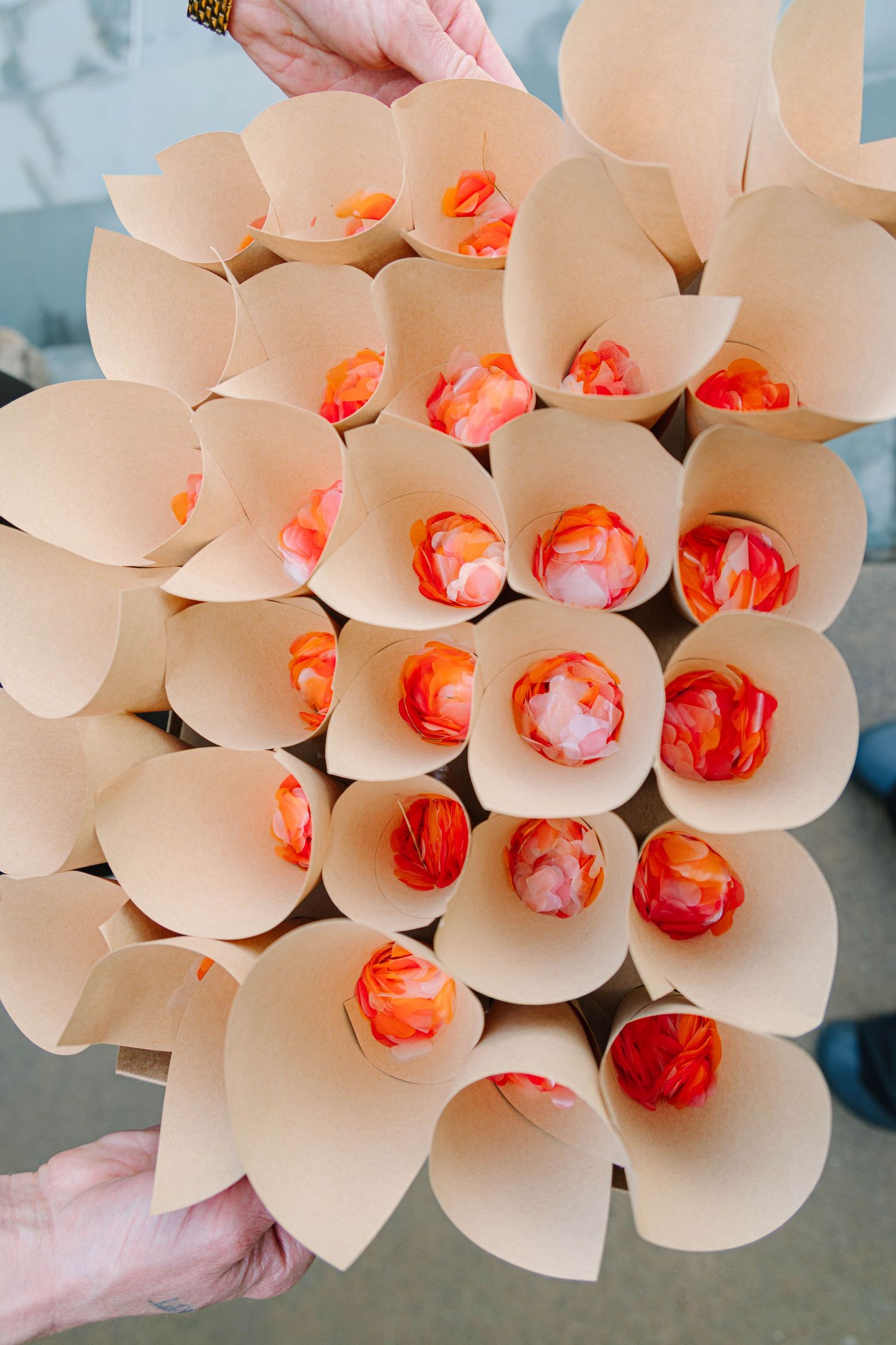 Hands holding tray of kraft paper cones filled with orange and pink flower petals for wedding toss.