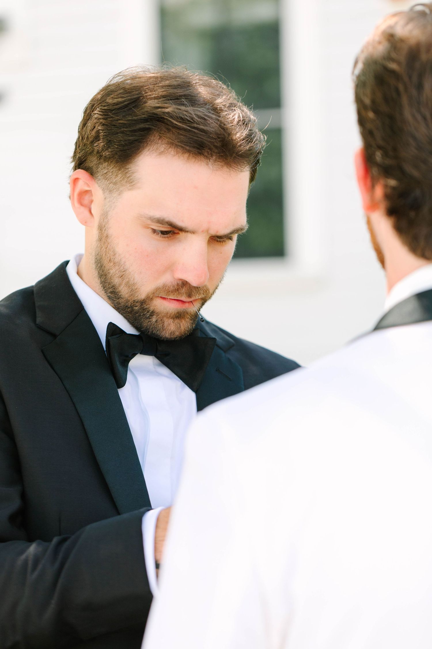 Groomsman in black tuxedo looking down while adjusting another mans cufflinks outdoors.