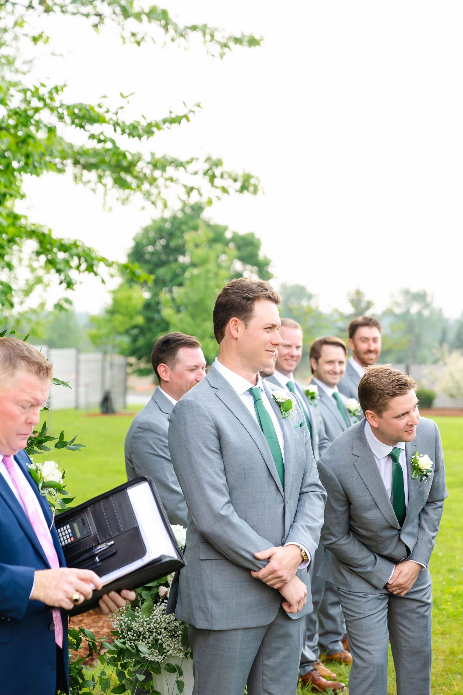 Groom and groomsmen in matching gray suits with green ties standing at outdoor wedding ceremony.