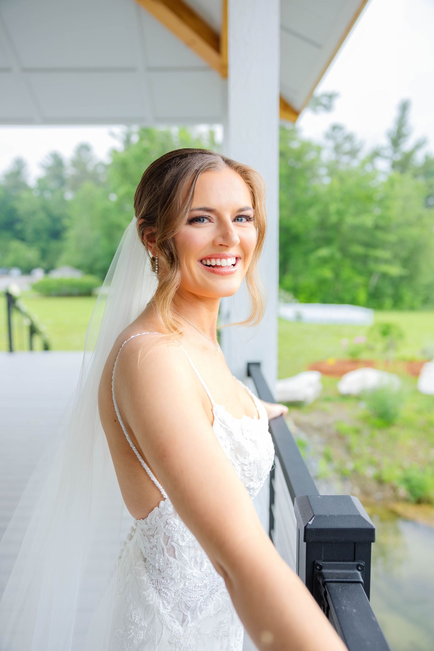 Beautiful bride in lace spaghetti strap wedding gown smiles on venue balcony with green backdrop.