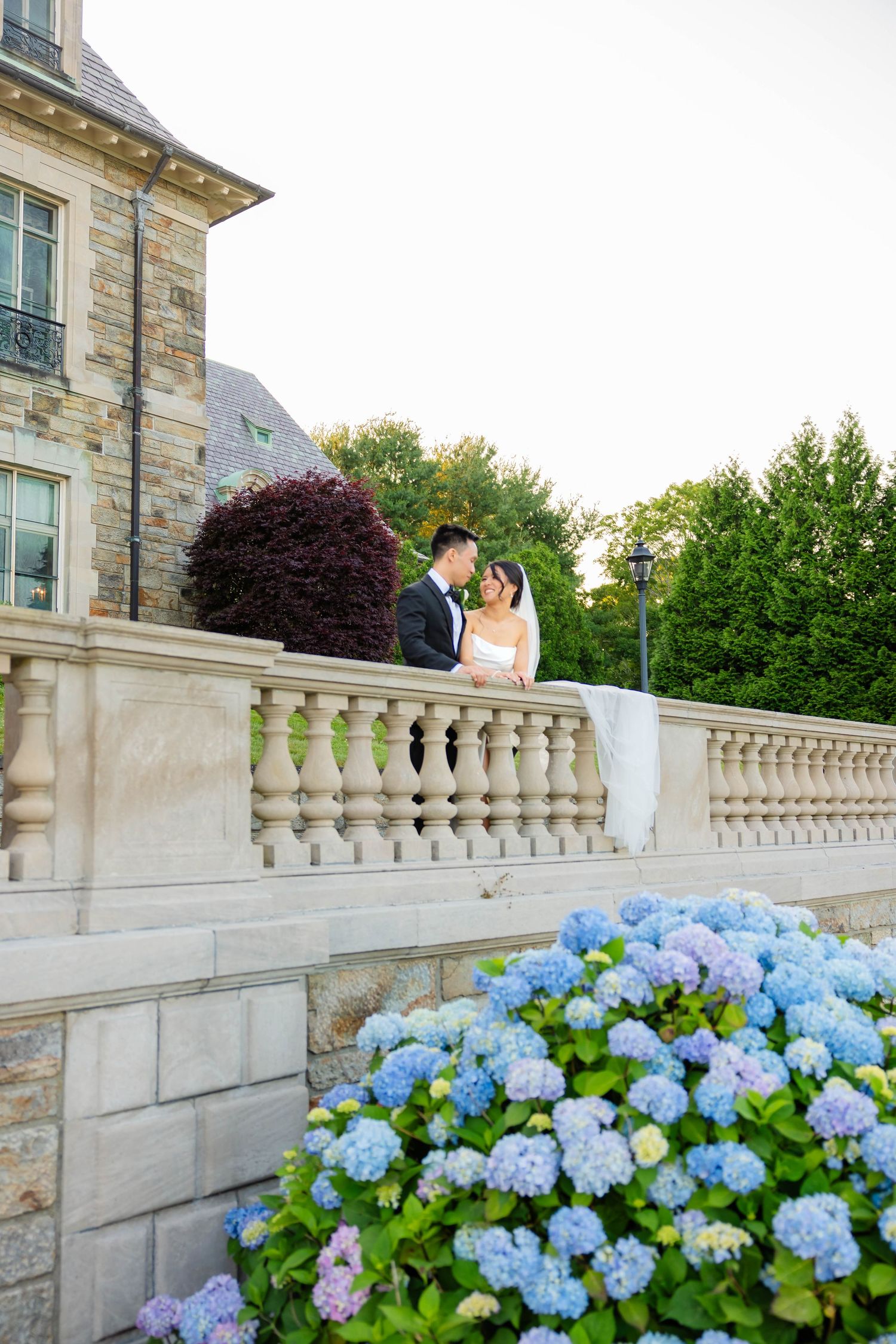 A wedding couple shares a romantic moment on a stone balustrade terrace surrounded by blue hydrangeas and lush gardens.