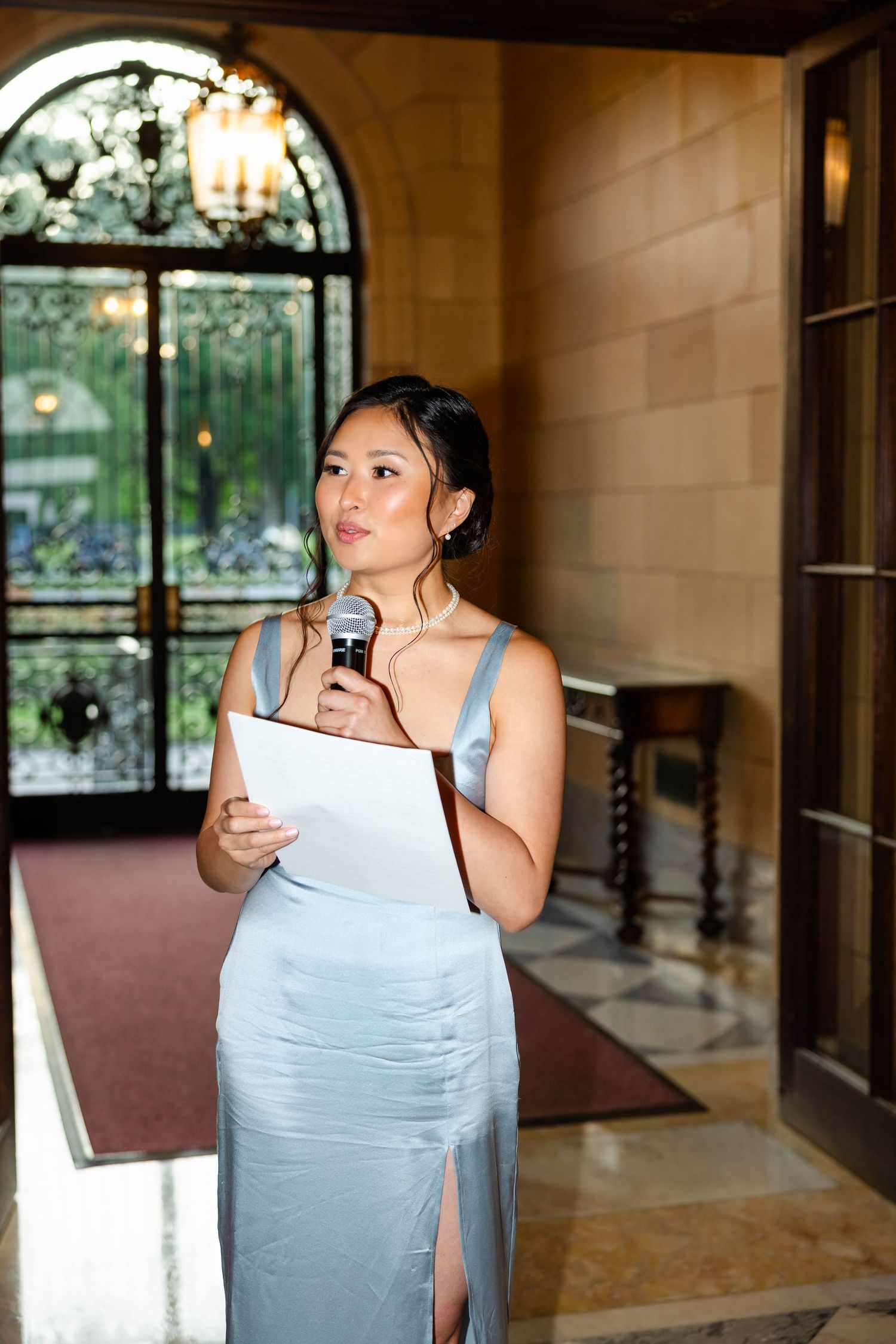 Woman in silver gown holding microphone and papers giving speech in elegant venue with ornate iron doors.