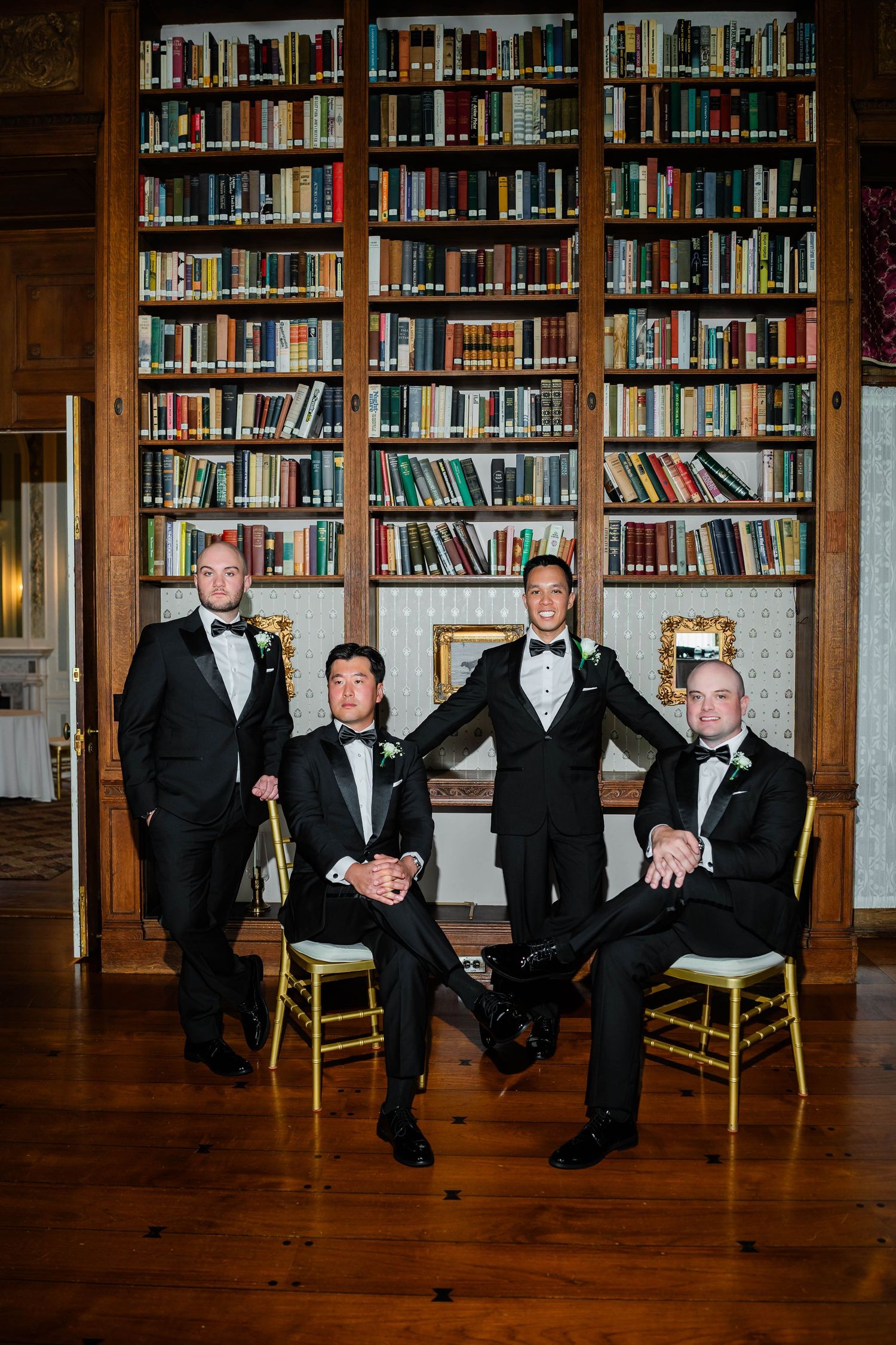 Four groomsmen in black tuxedos posing in front of a large bookshelf library at a wedding.