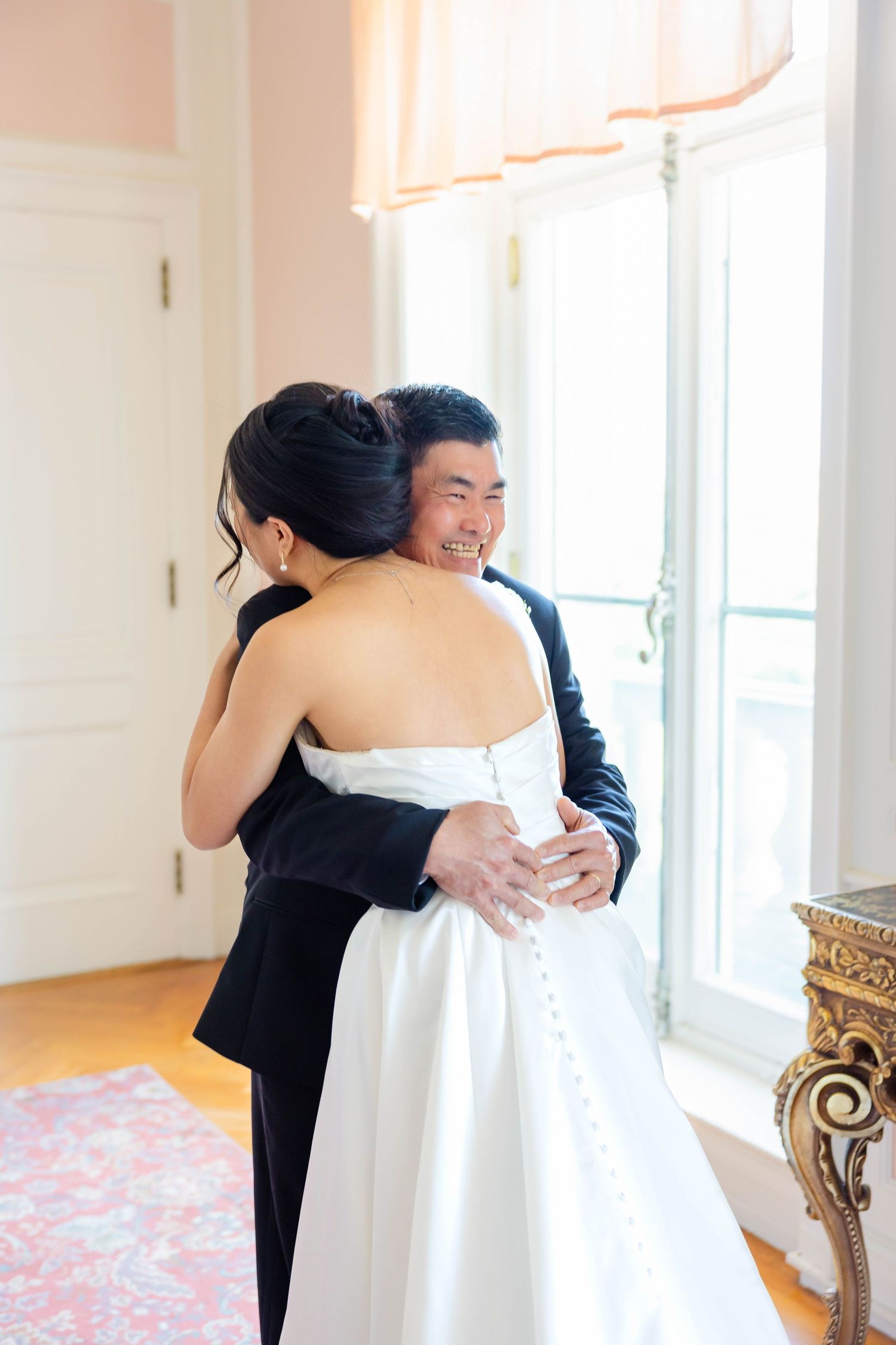 Smiling groom in black tuxedo embracing bride in white strapless wedding gown during emotional first look indoors.
