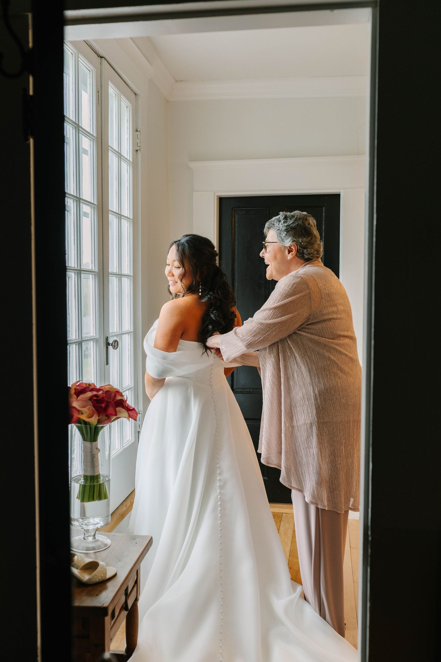 Mother helping bride in off-shoulder white gown with dress buttons in bright windowed room.
