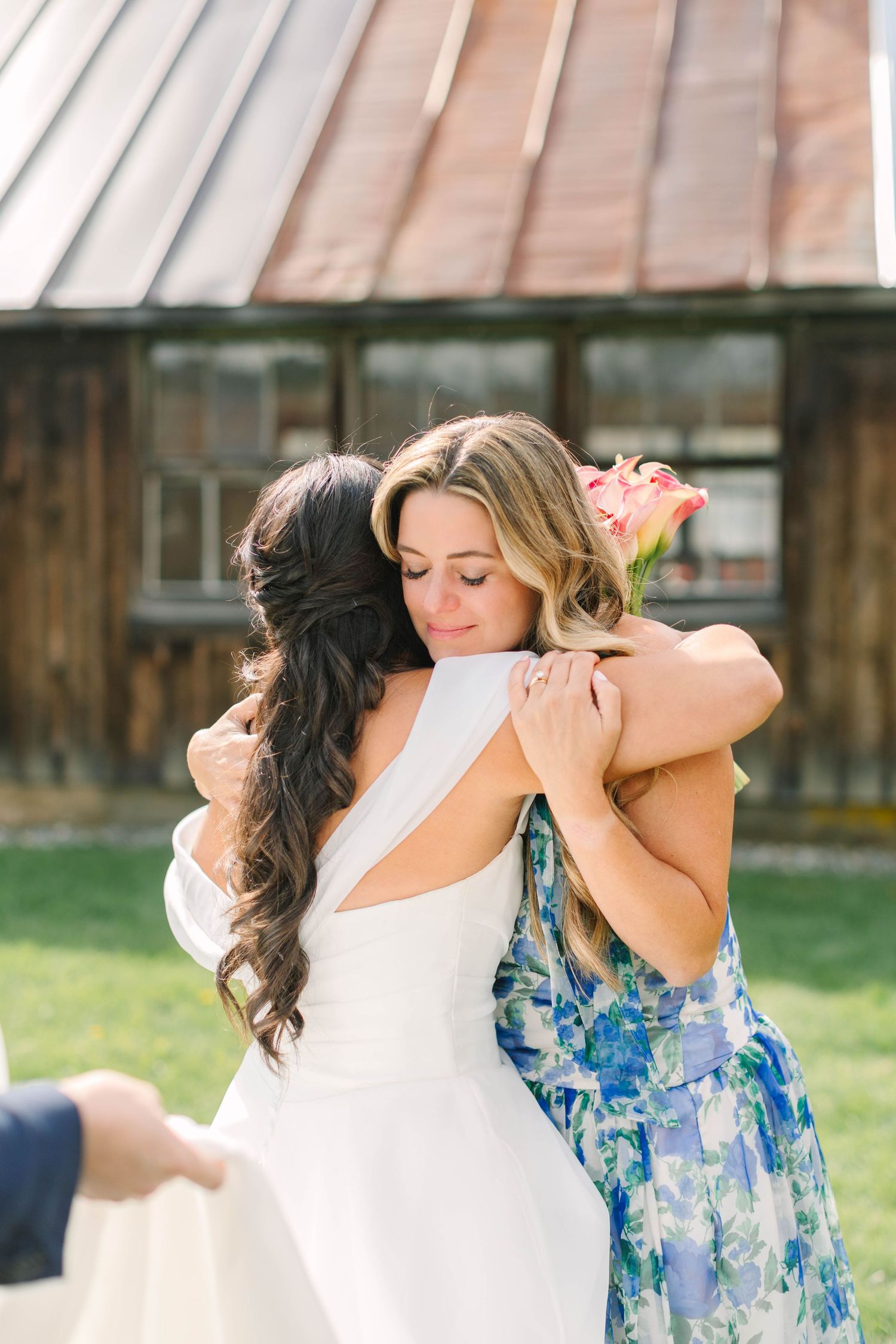 Bride in white gown shares emotional hug with guest in floral dress holding pink rose at outdoor wedding venue.