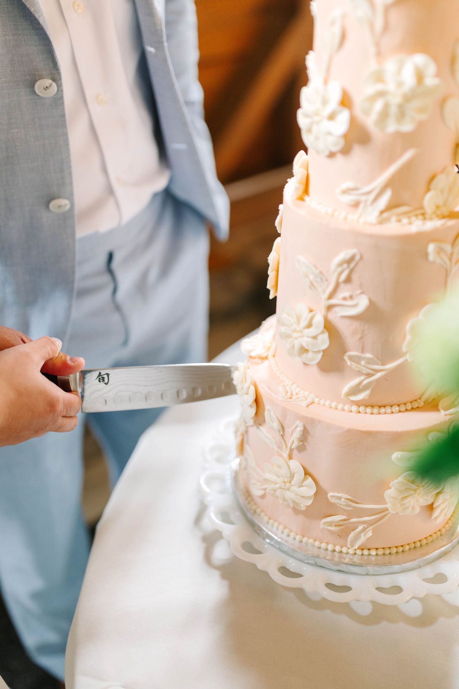 Groom in blue suit cutting a three-tier pink wedding cake with white floral decorations.