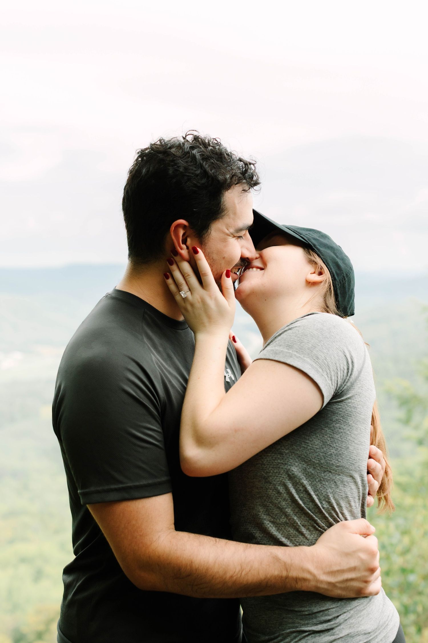 A couple shares a romantic embrace and kiss outdoors with a scenic green mountain landscape in the background.