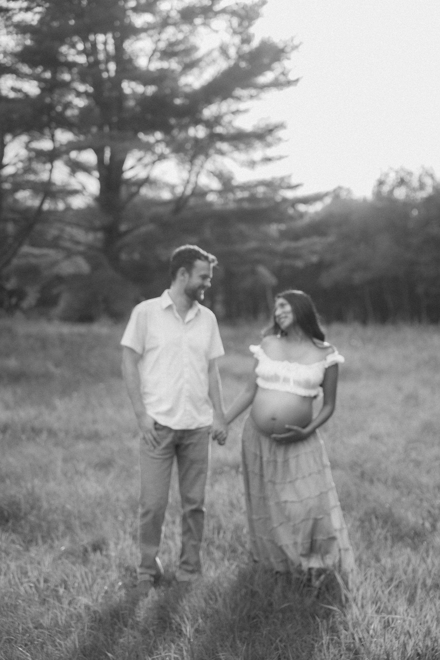 Black and white maternity photo of a couple holding hands in a field, smiling at each other at sunset.