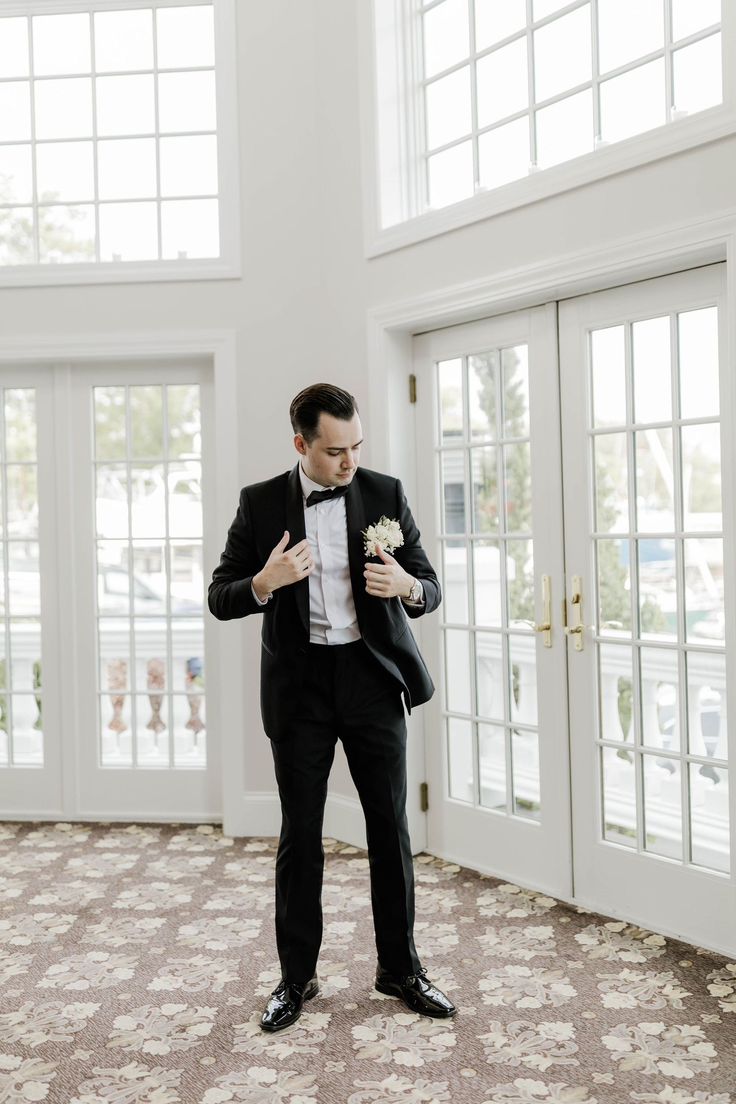Groom in black tuxedo adjusting white boutonniere indoors near French doors.