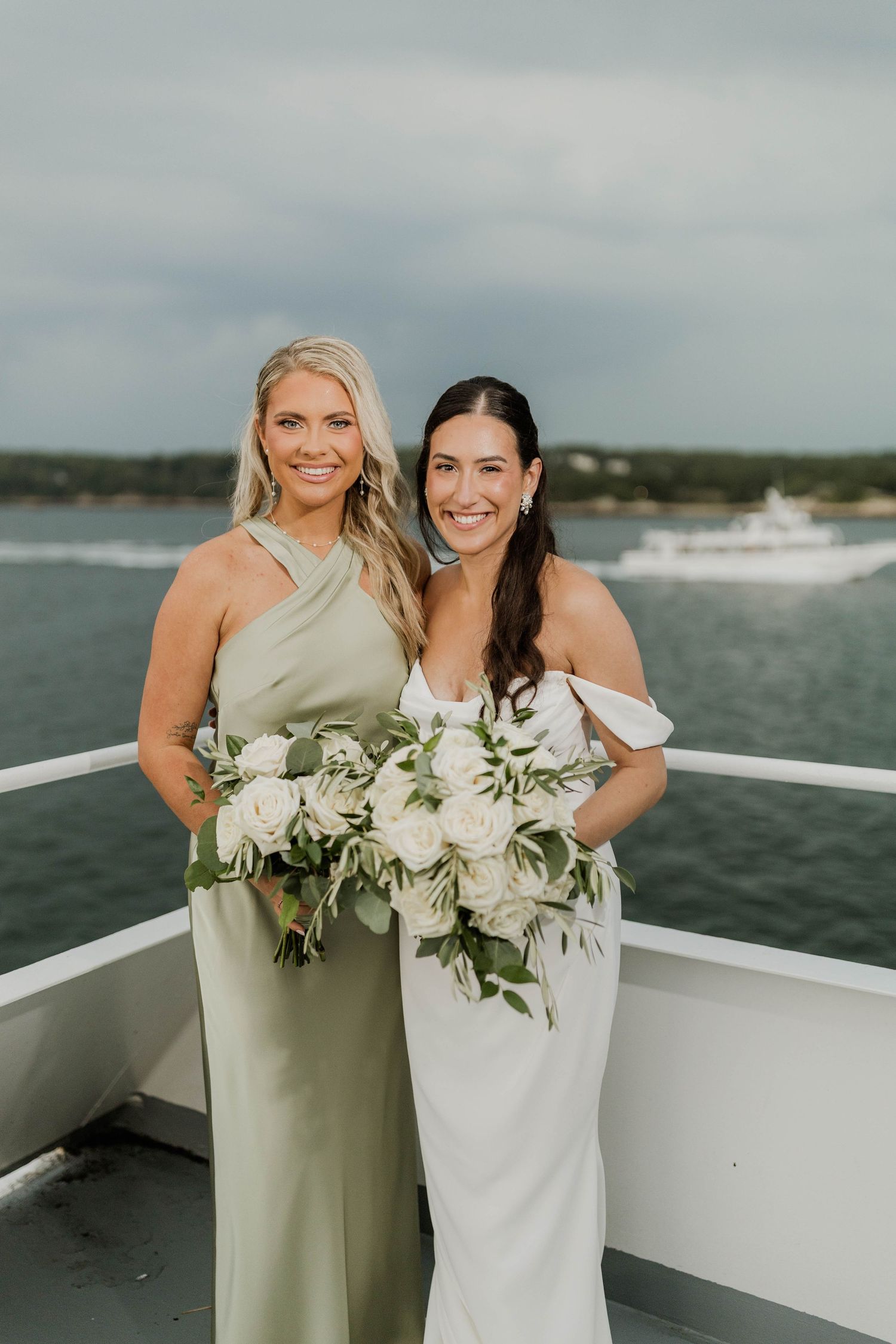 Bride in white off-shoulder gown poses with bridesmaid in sage green halter dress on yacht deck.