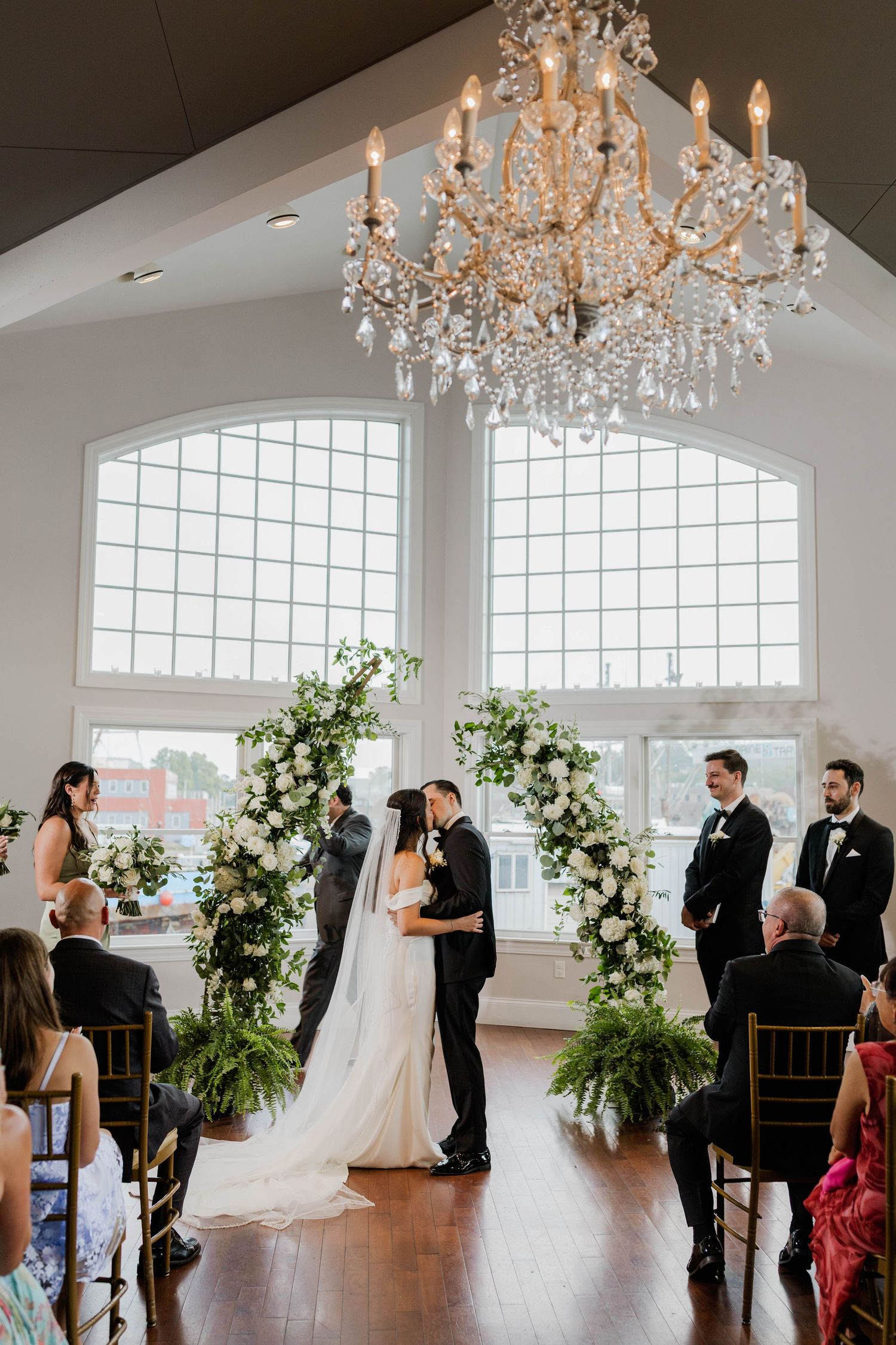 Bride and groom share first kiss at floral arch altar under crystal chandelier in elegant venue.
