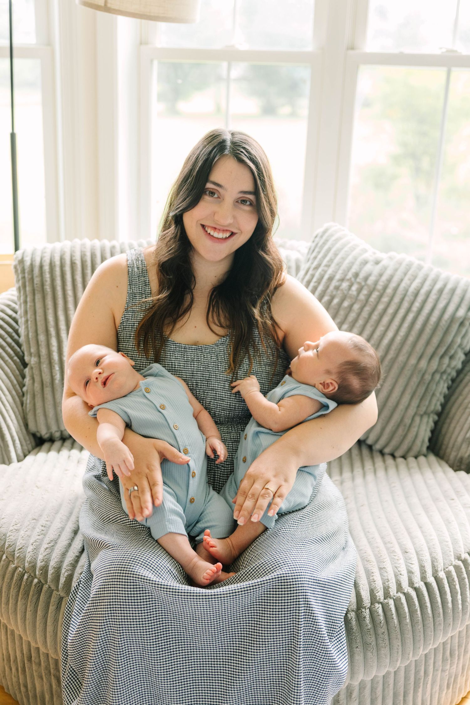 Smiling mother sitting on a cozy chair holding two newborn twins dressed in blue outfits near a bright window.