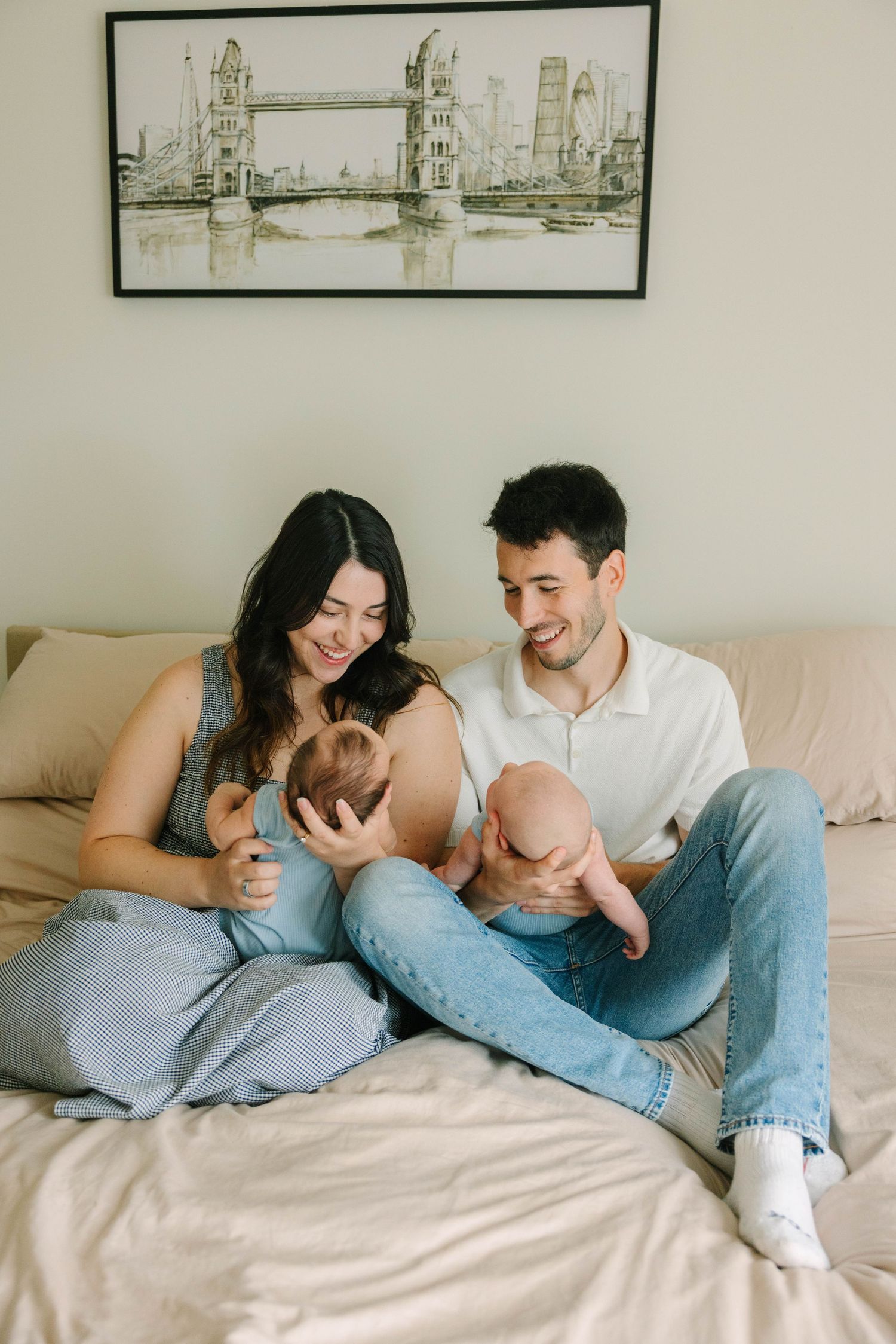 Happy parents sitting on a bed holding their twin newborns with a London bridge artwork above.