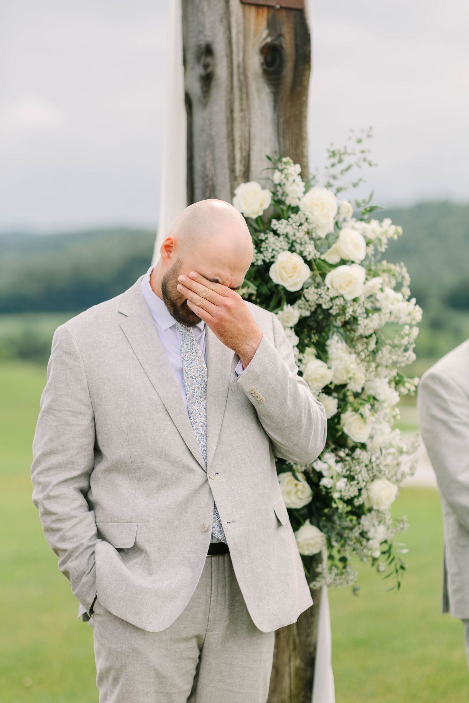 Emotional groom wipes tears beside a floral-decorated rustic wooden arch during outdoor wedding ceremony.