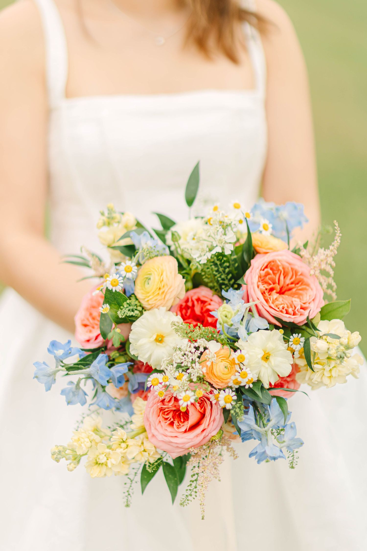 Bride holds a vibrant wedding bouquet featuring coral garden roses, blue delphiniums, daisies, and ranunculus blooms.