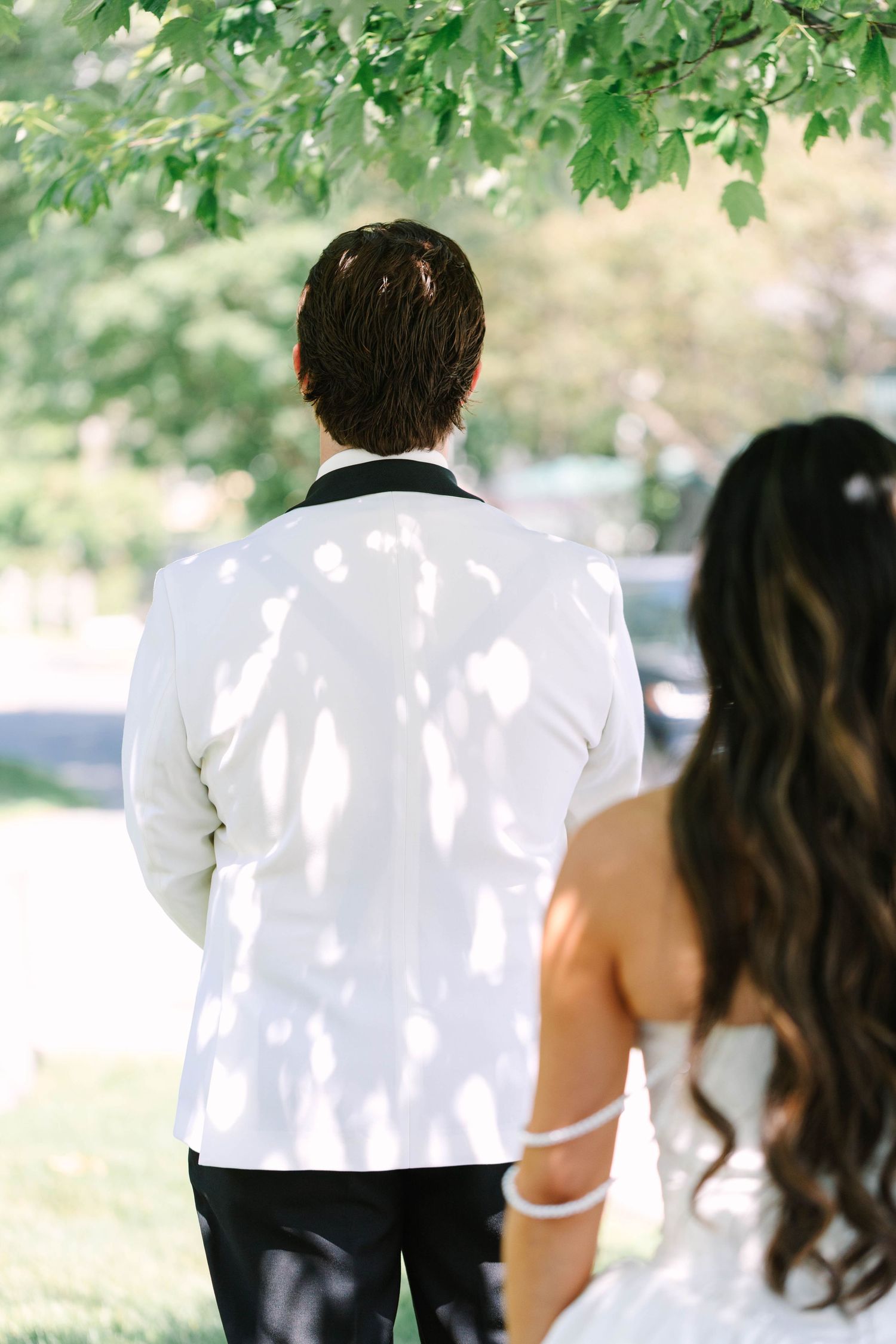 Groom in white tuxedo jacket stands with back turned during first look with bride outdoors.