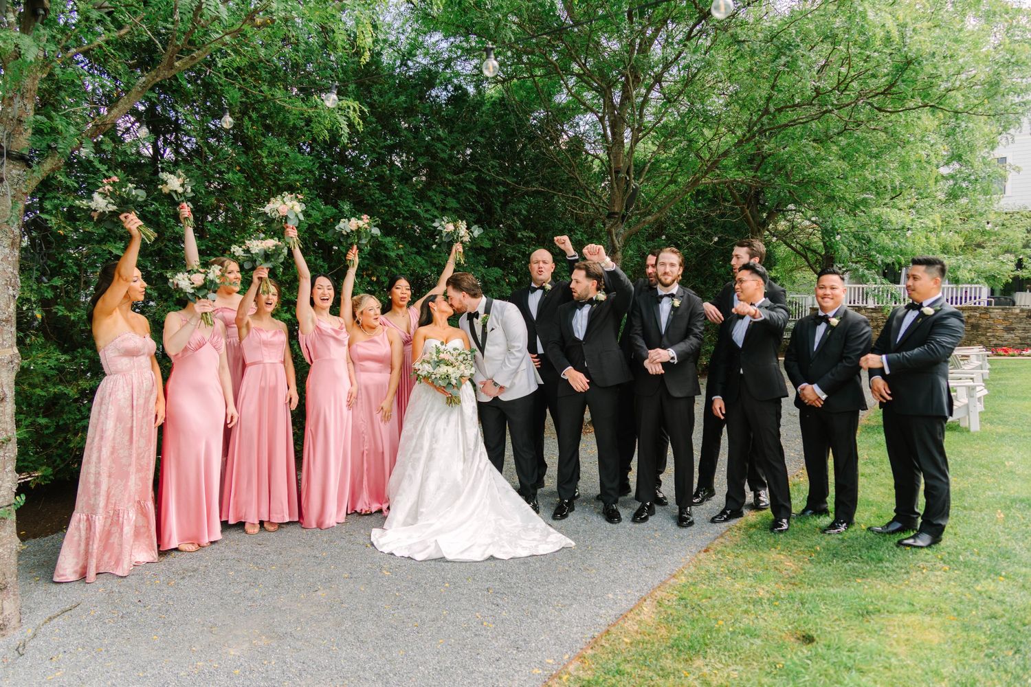 Joyful wedding party celebrating outdoors with bridesmaids raising bouquets and groomsmen in tuxedos.