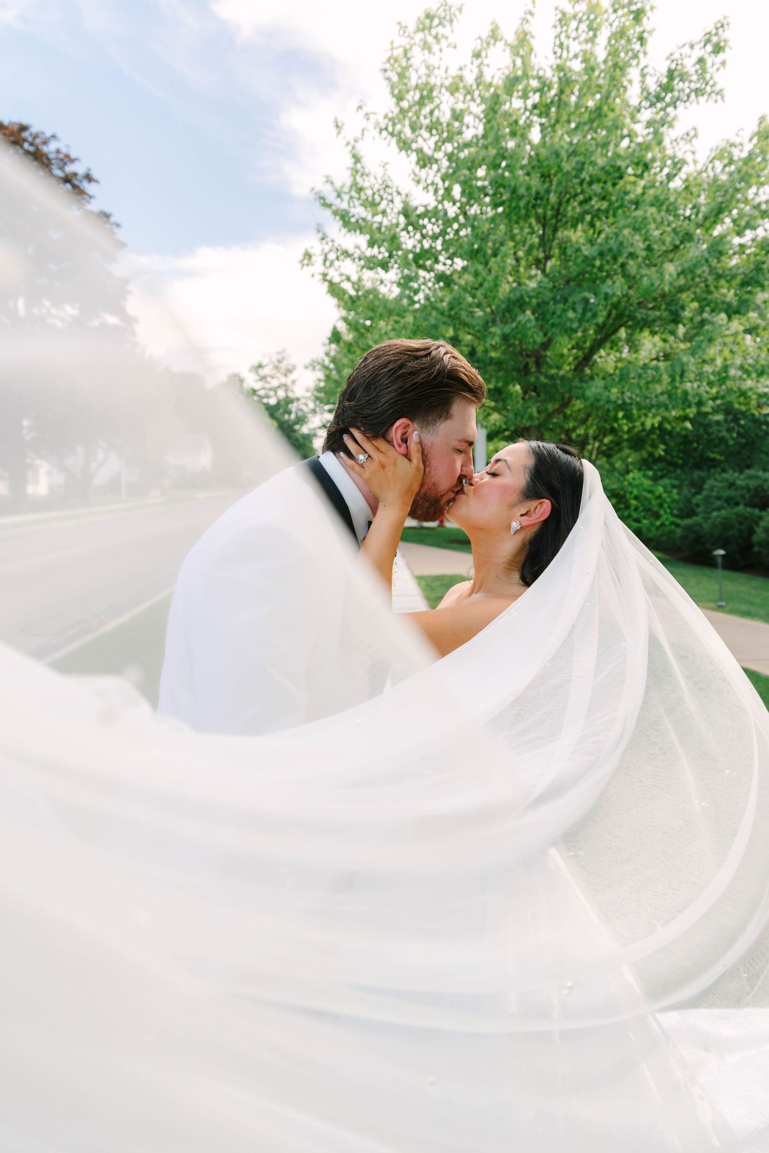 Bride and groom sharing a romantic kiss outdoors, wrapped in a flowing white wedding veil with green trees in background.
