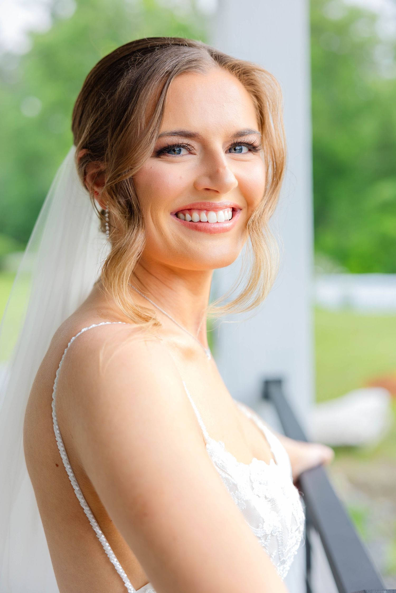 Close-up portrait of radiant bride in lace wedding gown with veil smiling on outdoor balcony.