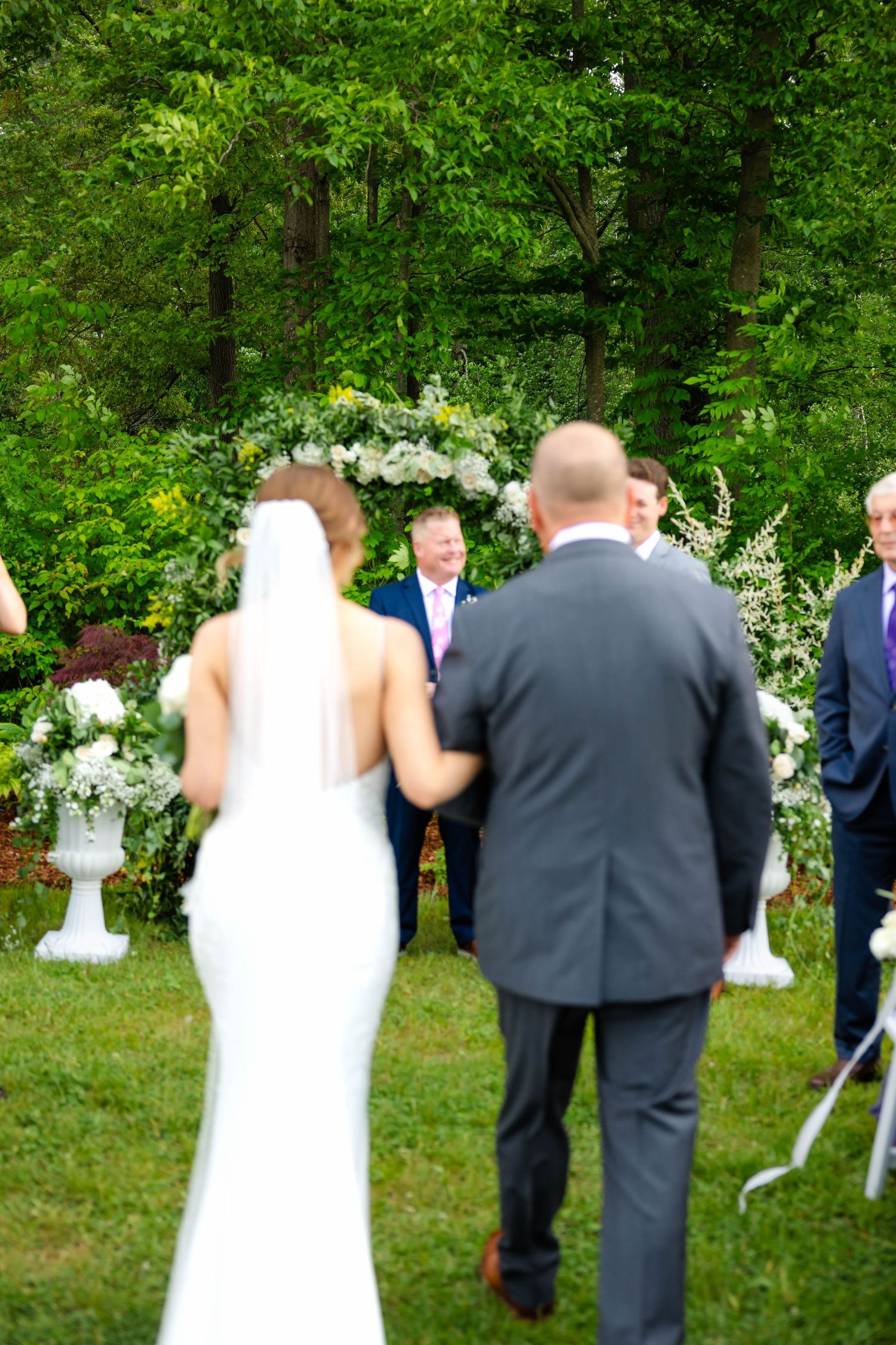 Bride walking down grassy aisle toward groom at outdoor ceremony with floral arch backdrop.