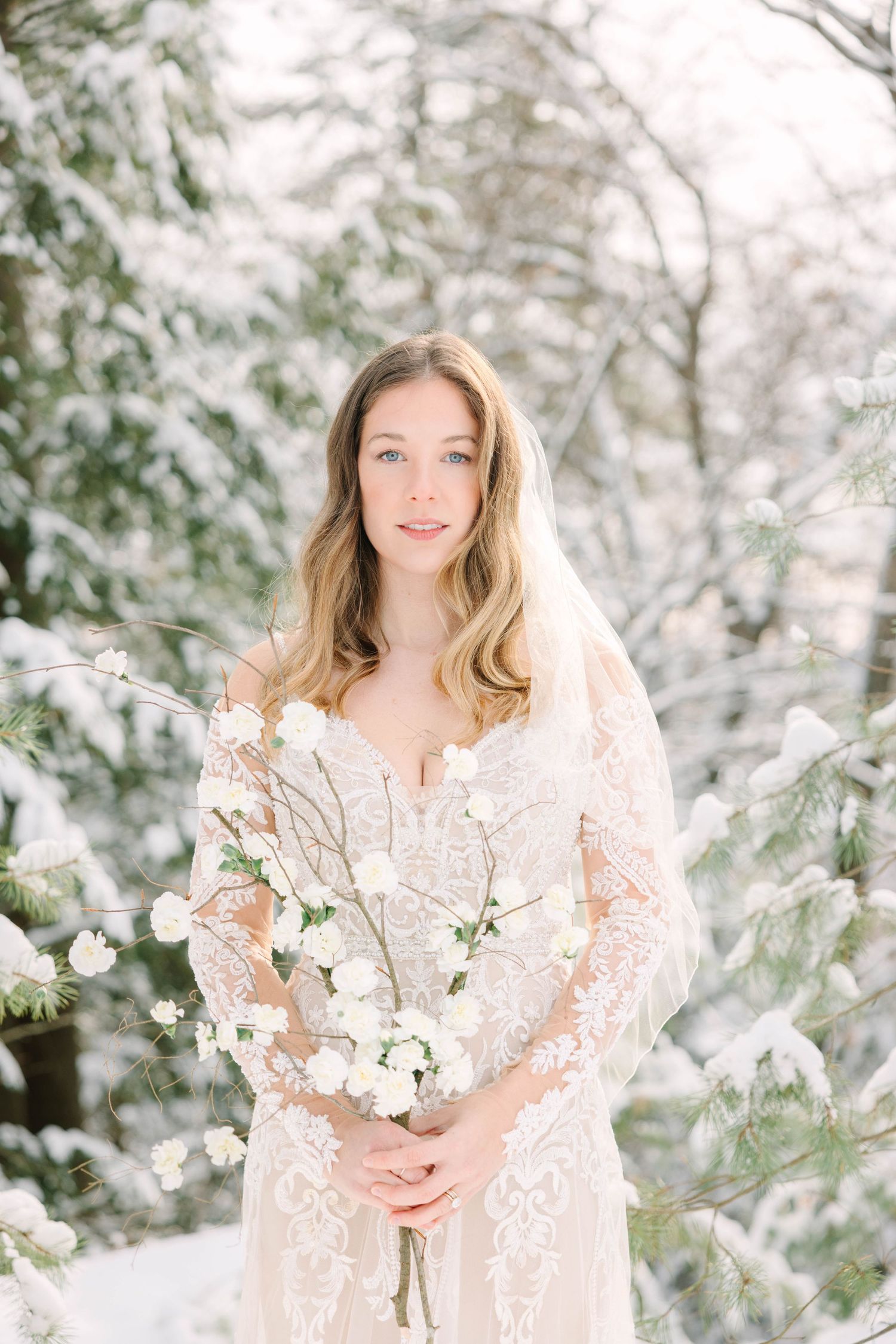 Bride in lace gown and veil holds white flowers in snowy winter forest setting.